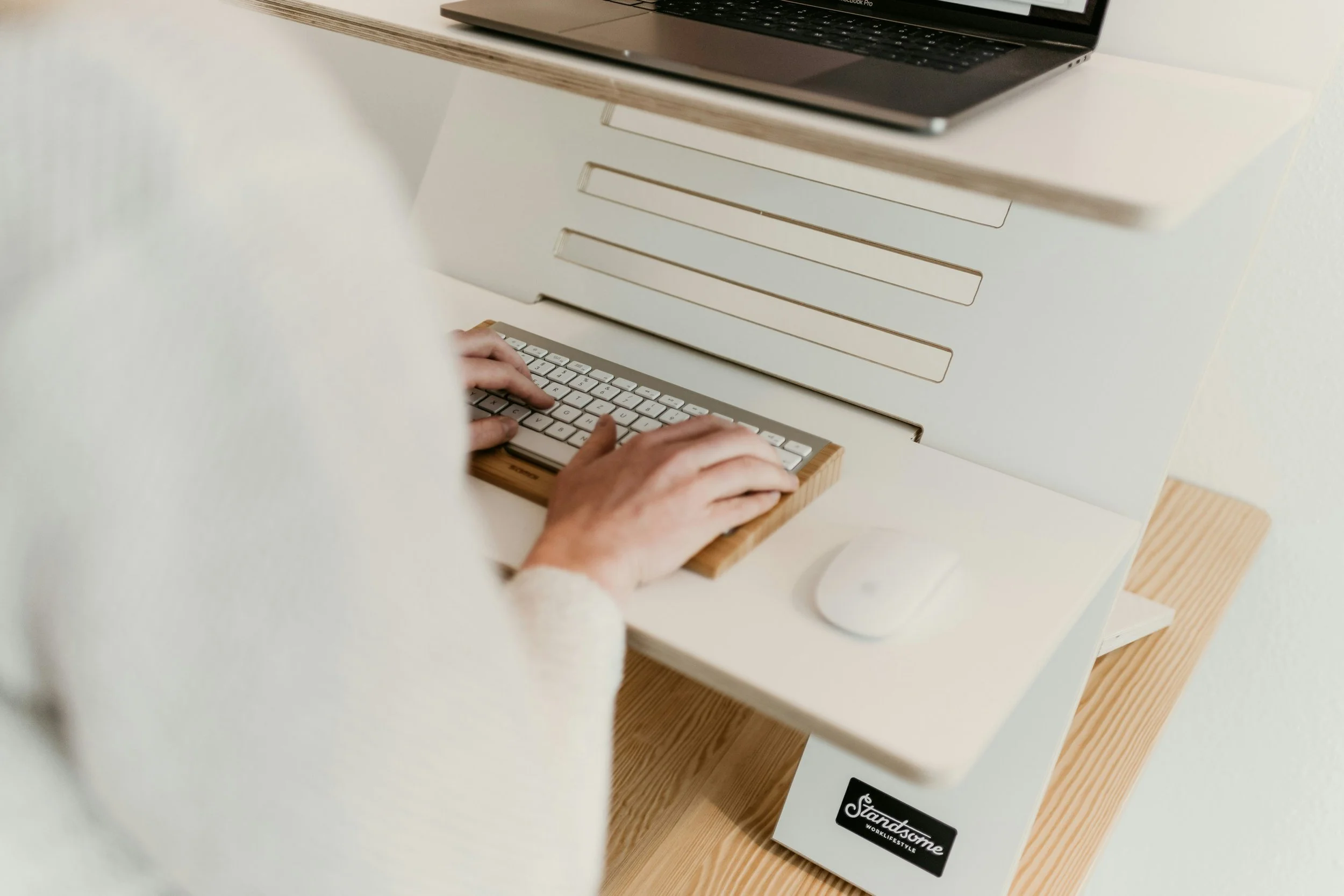 Person typing on a small keyboard on a minimalist white desk with a white mouse, a closed laptop on a wooden shelf, and a white wall behind.