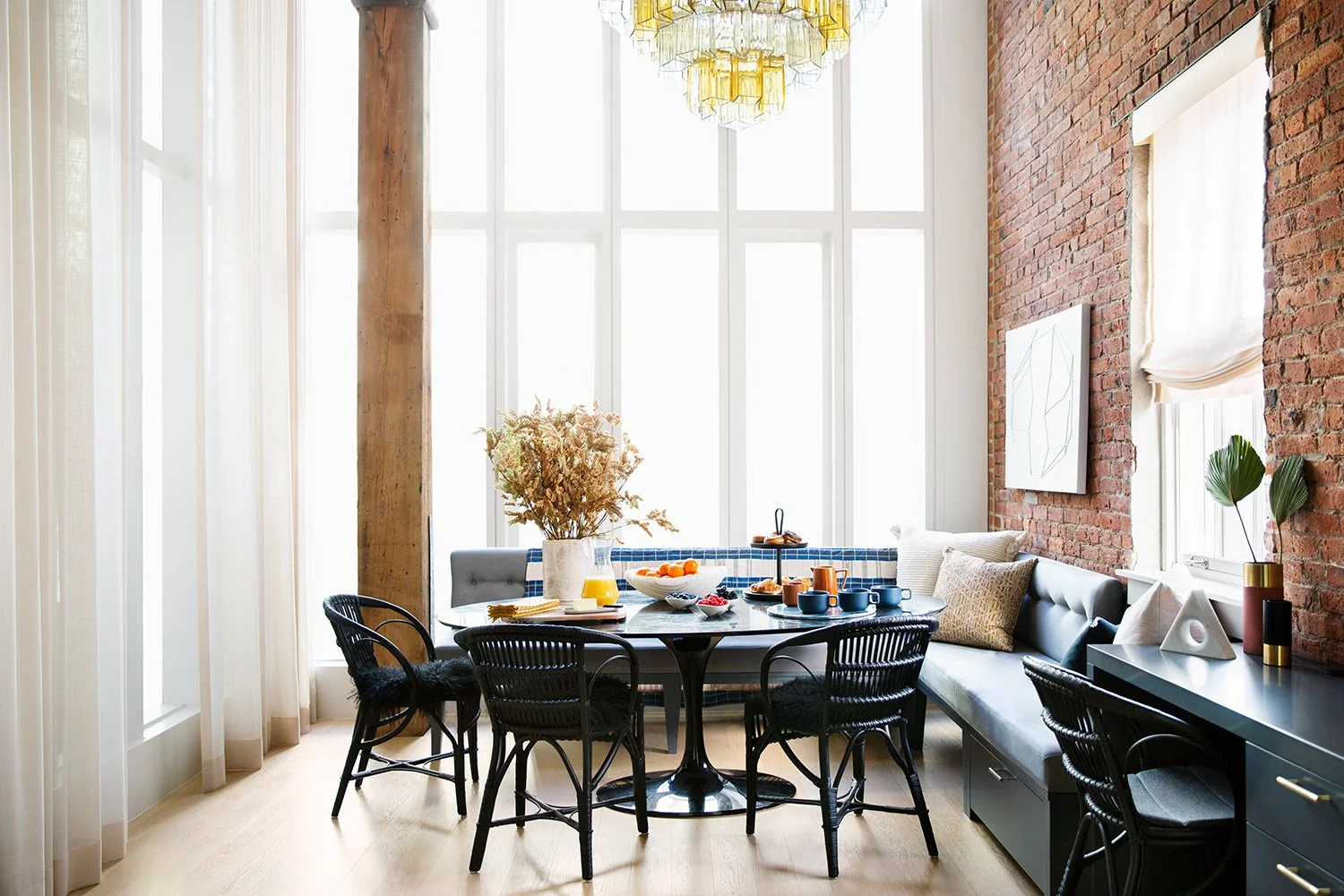 Dining area with a round table set for breakfast, black chairs, a built-in banquette, and decorative elements like vases, pillows, and artwork in a bright room with large windows and an exposed brick wall.