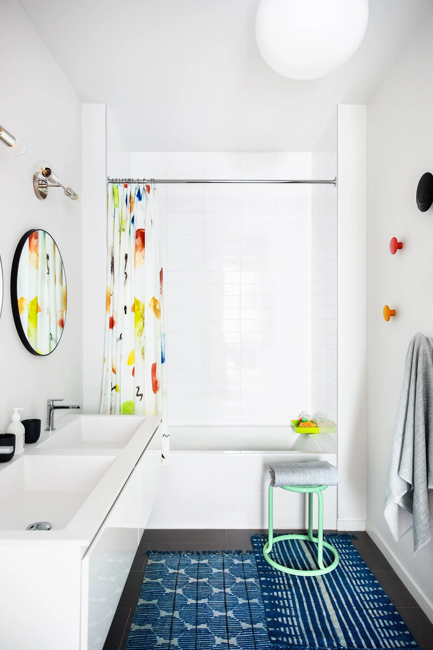 A bright, minimalist bathroom with white walls and ceiling. Features a white vanity with a black soap dispenser and cup, a round mirror, colorful hooks on the wall, and a shower area with a white tile wall and colorful curtain. A small stool with a g