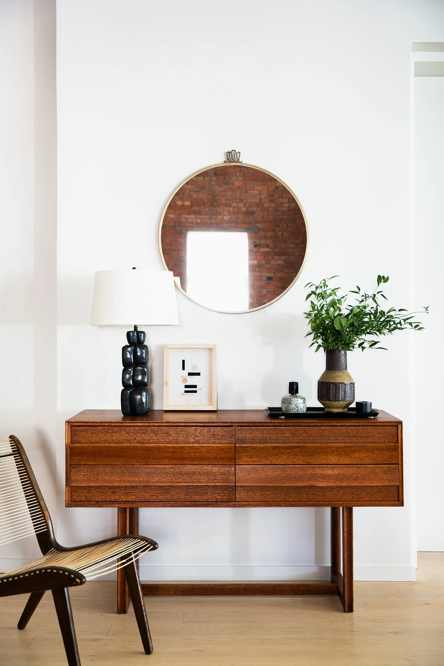 Interior of a modern entryway with a wooden sideboard, a large round mirror, a table lamp, a small framed artwork, a green plant in a decorative vase, and a small decorative bottle on a tray, against a white wall with a brick wall reflected in the mi