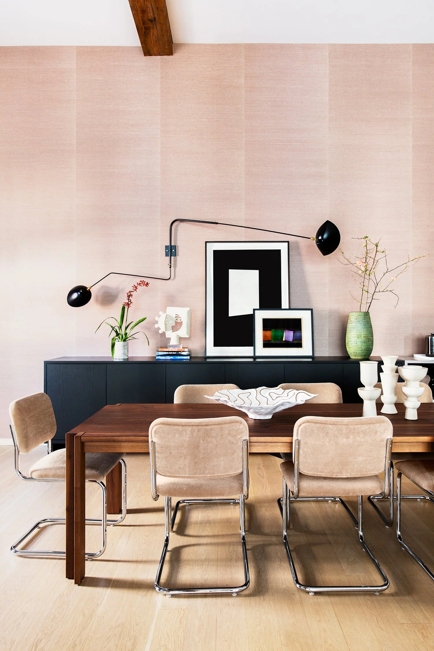 A modern dining room with a wooden table, beige upholstered chairs, and a black sideboard against a pink wall. The wall features black and white framed artwork, a decorative sculpture, and several vases with plants and flowers. A black, adjustable wa