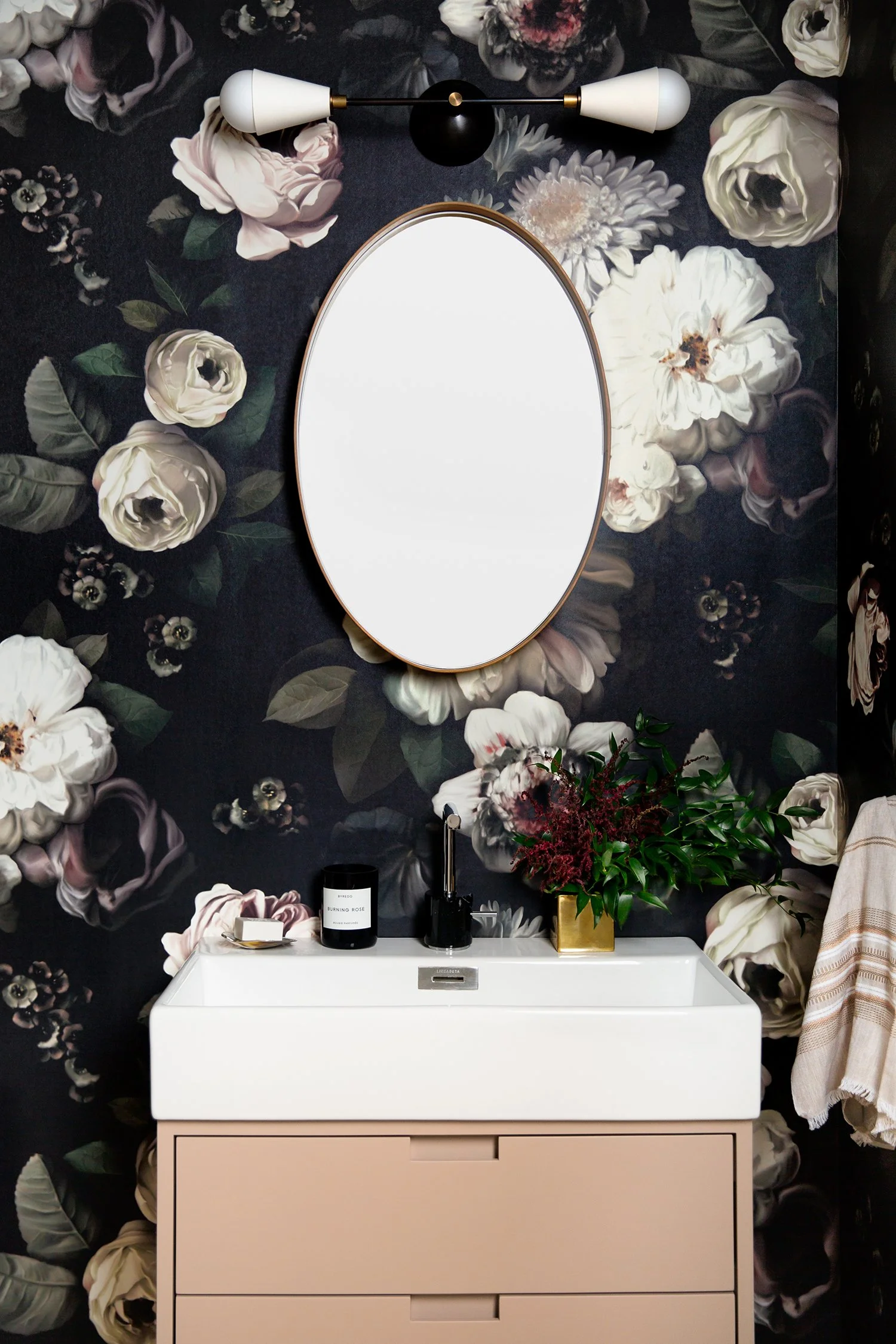 A modern bathroom vanity with a white sink, a gold vase with green leafy plants, candle, and toiletries, against a dark floral wallpaper with large white, pink, and purple flowers, and a round mirror above the sink.