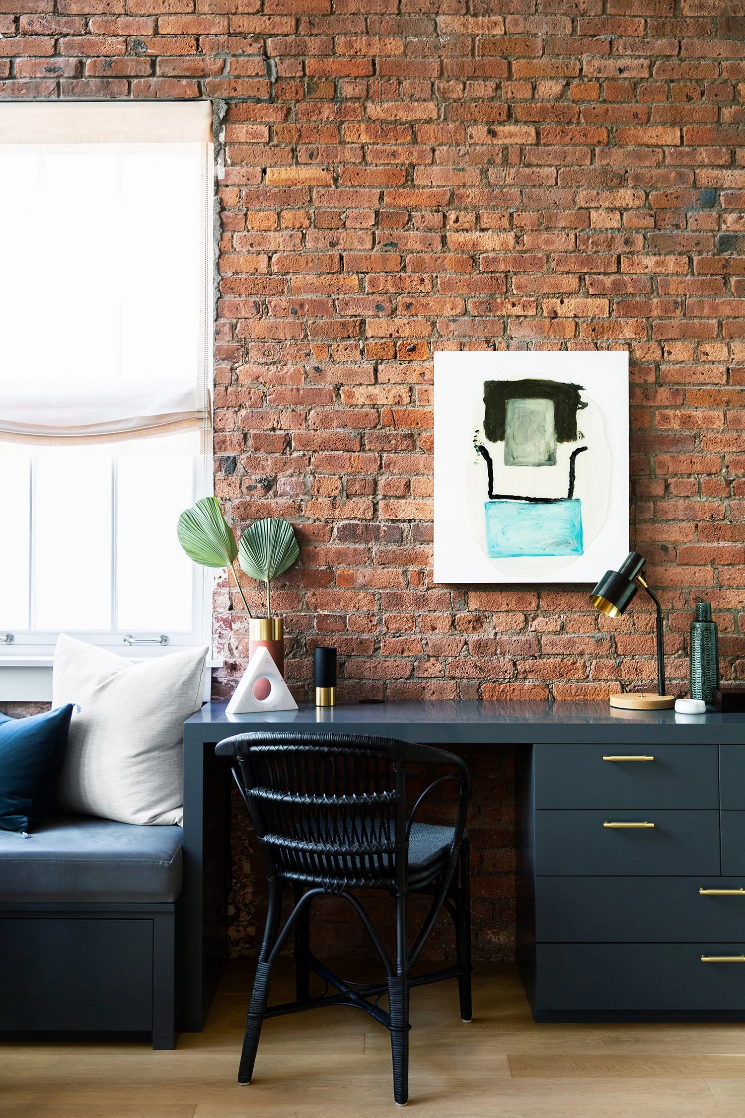 Modern workspace with a black desk and chair against an exposed brick wall, decorated with abstract art, a table lamp, and decorative vases, next to a window with white blinds.