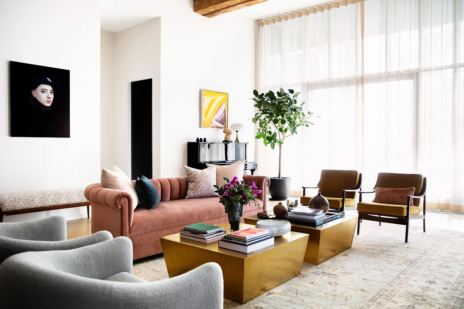 A modern living room with a pink velvet sofa, two brown armchairs, a large potted plant, a coffee table with books and decorative items, a white armchair, and artwork on white walls, all with large windows and sheer curtains.