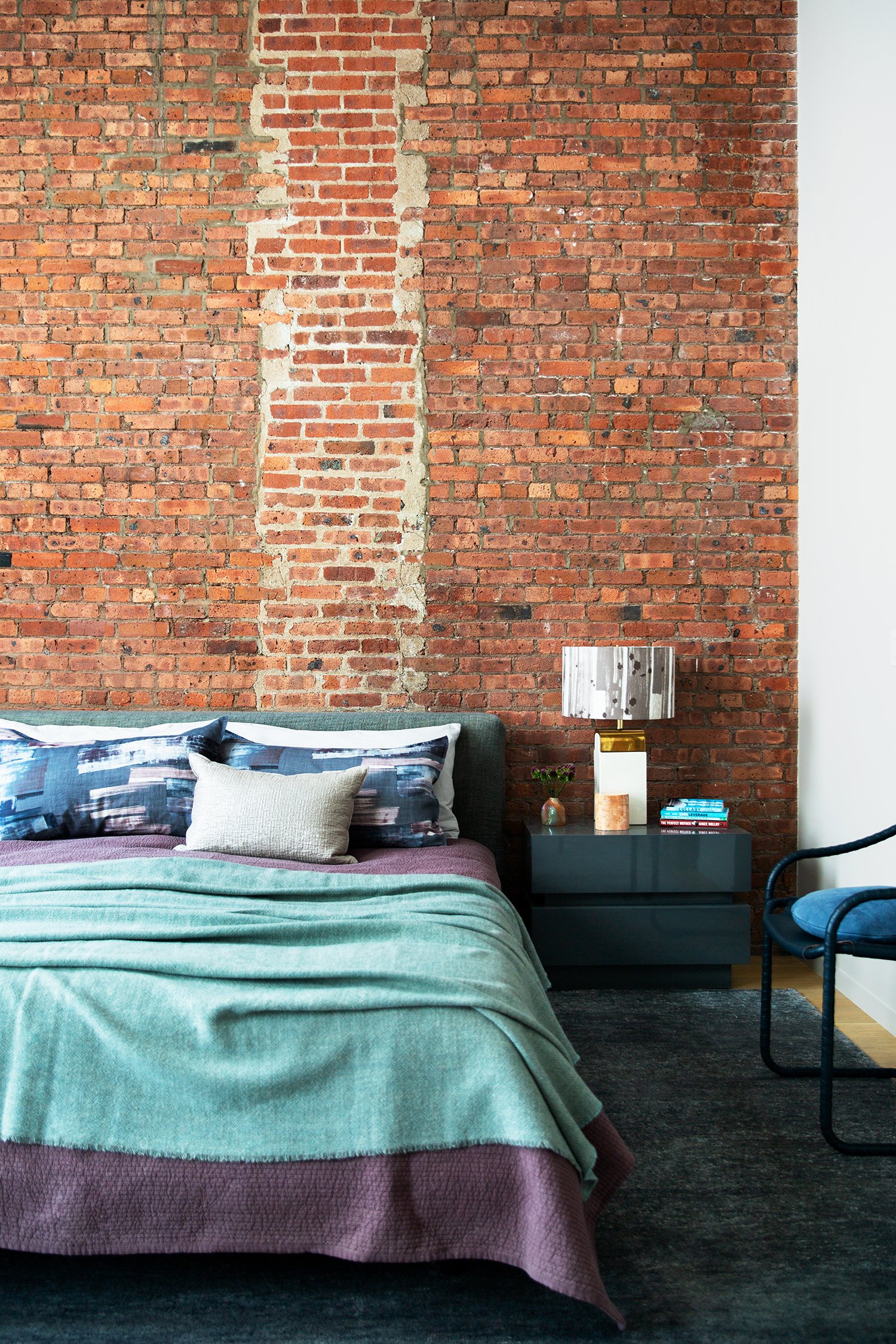 Bricked bedroom wall with a dark blue nightstand, a striped lamp, a small vase, and an assortment of books, beside a bed with green, purple, and patterned pillows.