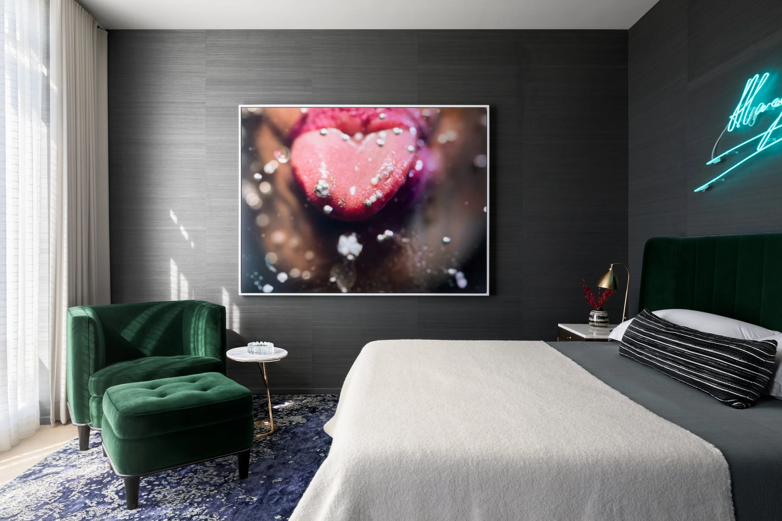 Modern bedroom with a dark gray wall, large framed close-up photo of a pink heart-shaped object with beads on it, green velvet chair and ottoman, white bed with dark gray and black pillows, side table with vase and lamp, and neon sign on the wall.