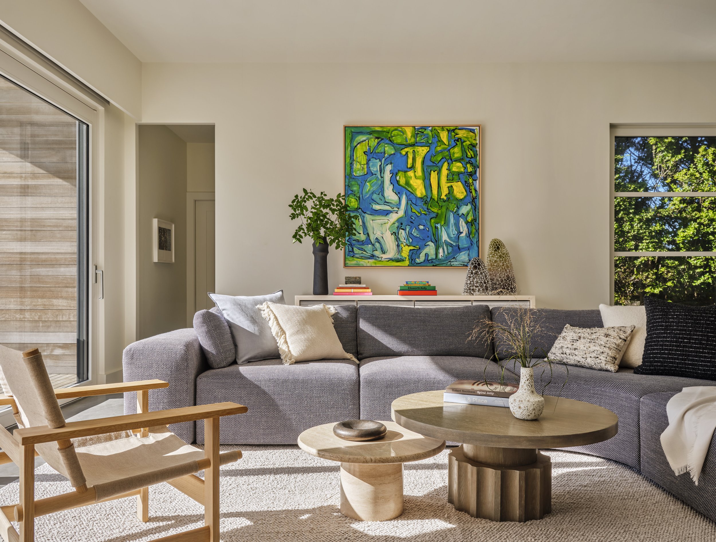Living room with a gray sectional sofa, wooden coffee tables, abstract artwork, a plant, and decorative objects, brightened by large windows showing greenery outside.