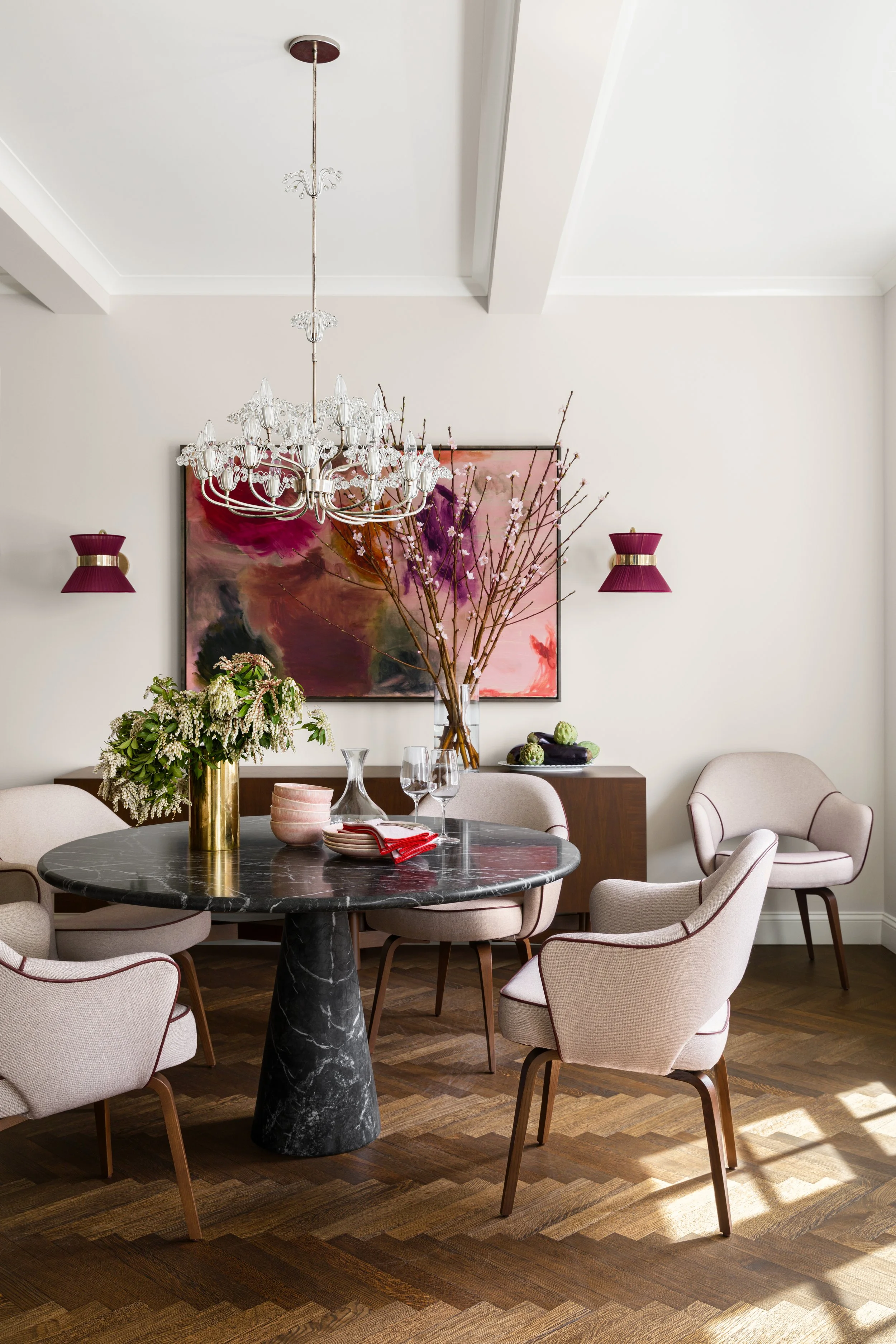 A modern dining room with a round black marble table, surrounded by five beige upholstered chairs with wooden legs. On the table are decorative dishes, glassware, and a red napkin. A tall gold vase holds branches with pink blossoms, and a small sideboard with styled decor is in the background. The wall features two purple sconces, and a large abstract painting with pink, purple, and black tones hangs on the wall. A crystal chandelier hangs from the ceiling, illuminating the room.