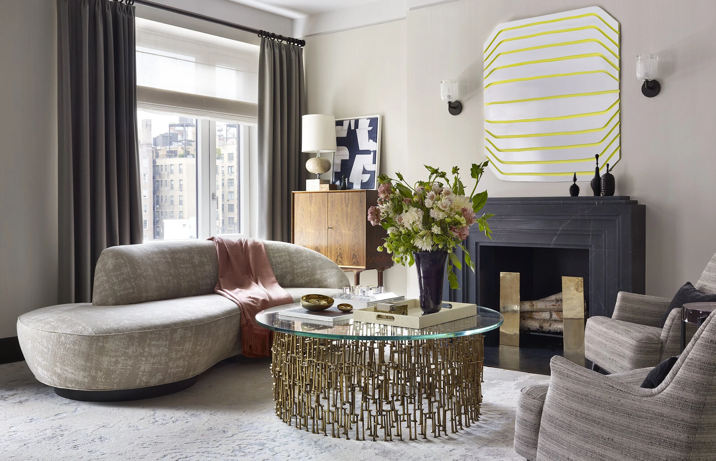 Living room with large window, beige curved sofa with pink throw, glass coffee table with gold base, flower arrangement, fireplace, modern wall art, and armchairs.
