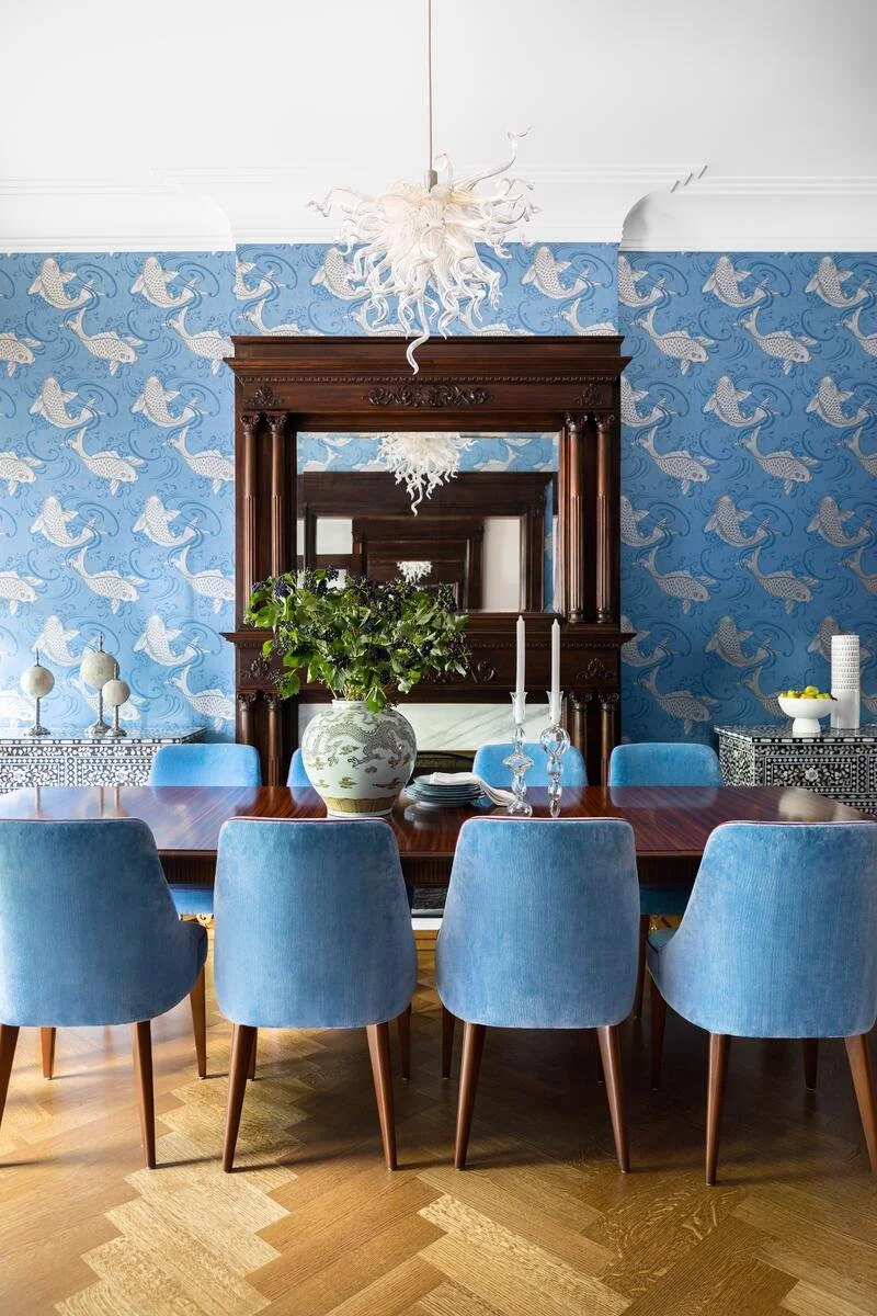 A dining room with a large wooden table surrounded by eight light blue chairs, a decorative mirror, a floral chandelier, a vase with green leaves, candles, and various decorative items on side tables, with wallpaper featuring white whales on blue waves.