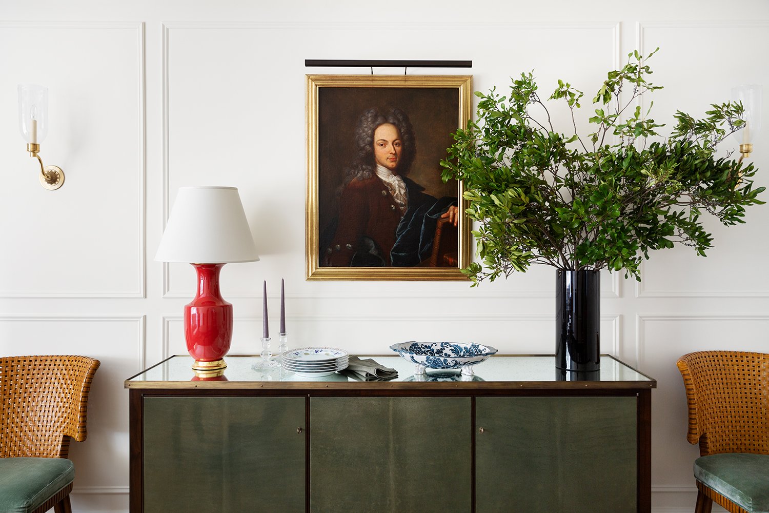 Interior of a room with white paneled walls, a large potted green plant in a black vase, a wooden sideboard with decorative dishes and candles, a red table lamp with a white shade, a framed portrait of a man with curly hair, and two wicker chairs with green cushions.