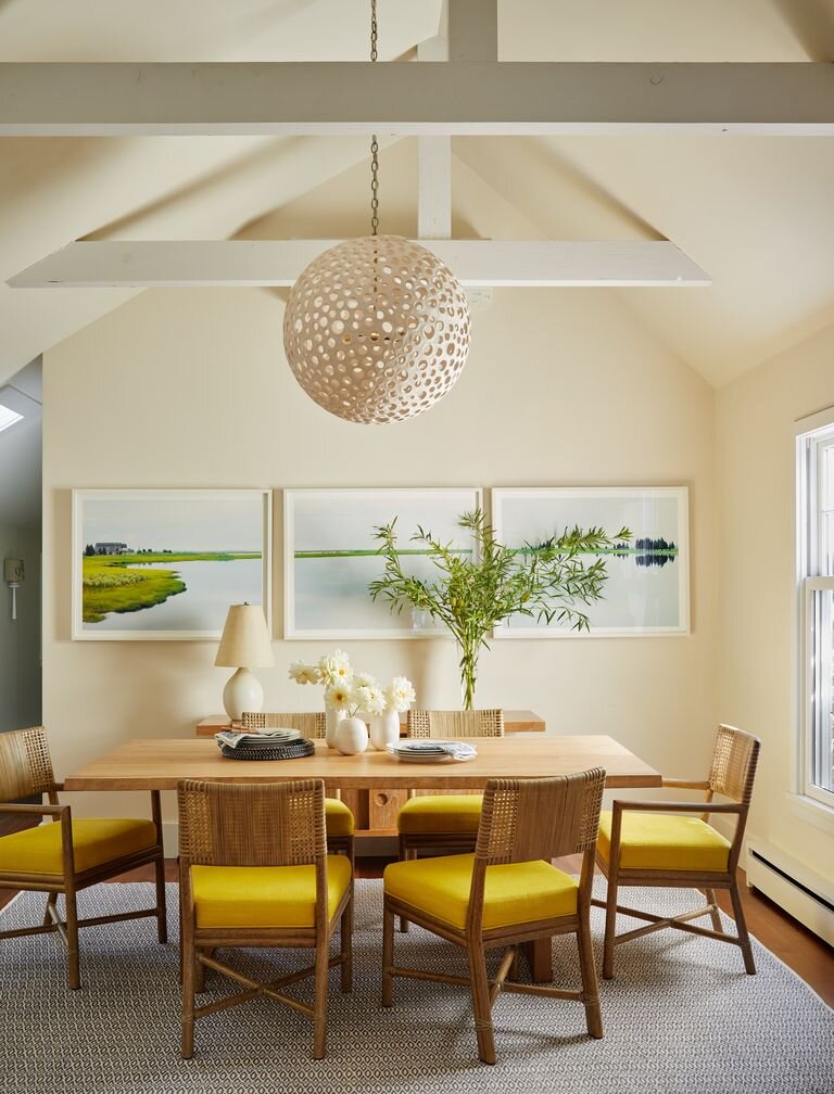 Dining room with wooden table, six chairs with yellow cushions, a large green plant, a lamp, and three framed landscape pictures on the wall.