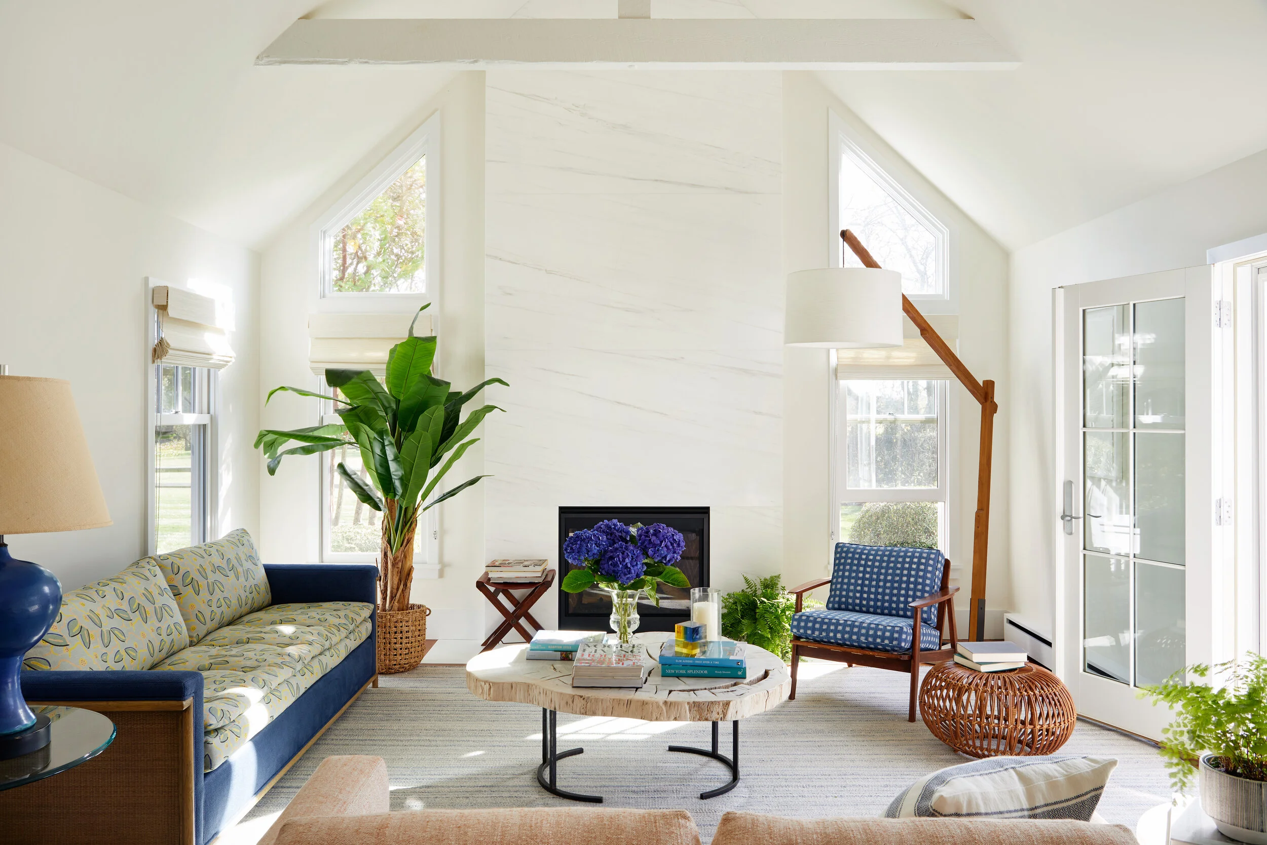 Bright living room with white walls, a white fireplace, large windows, a blue and yellow patterned sofa, a wooden armchair with blue upholstery, a large green potted plant, a round wooden coffee table with books and a vase of purple hydrangeas, a wic
