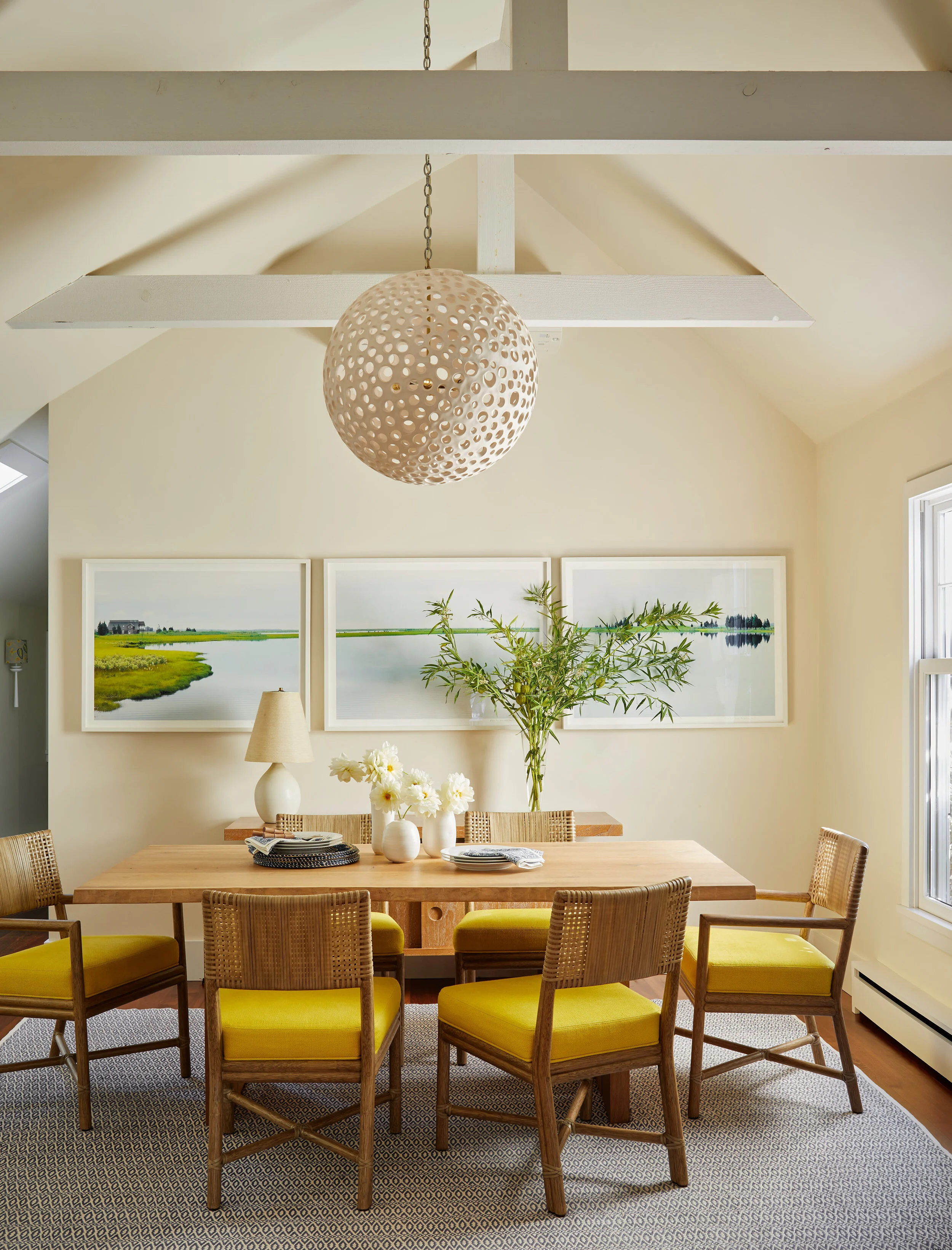 Dining room with wooden table, six yellow upholstered chairs, a large vase with greenery, framed landscape art, and a hanging spherical light fixture.