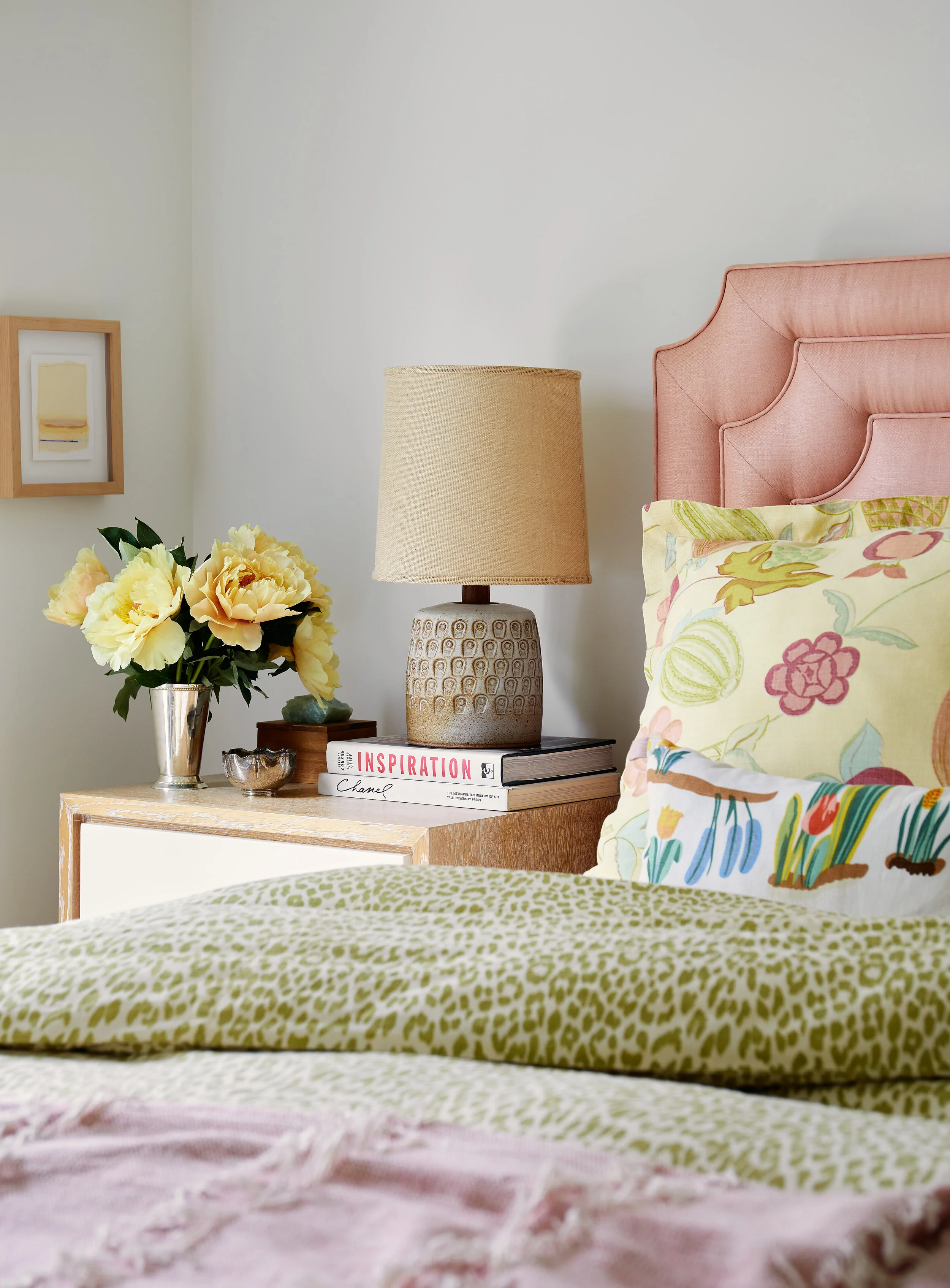 A close-up of a bedroom nightstand with a beige textured lamp, a vase of yellow flowers, and books, with a partial view of a pink upholstered headboard and colorful bedding.