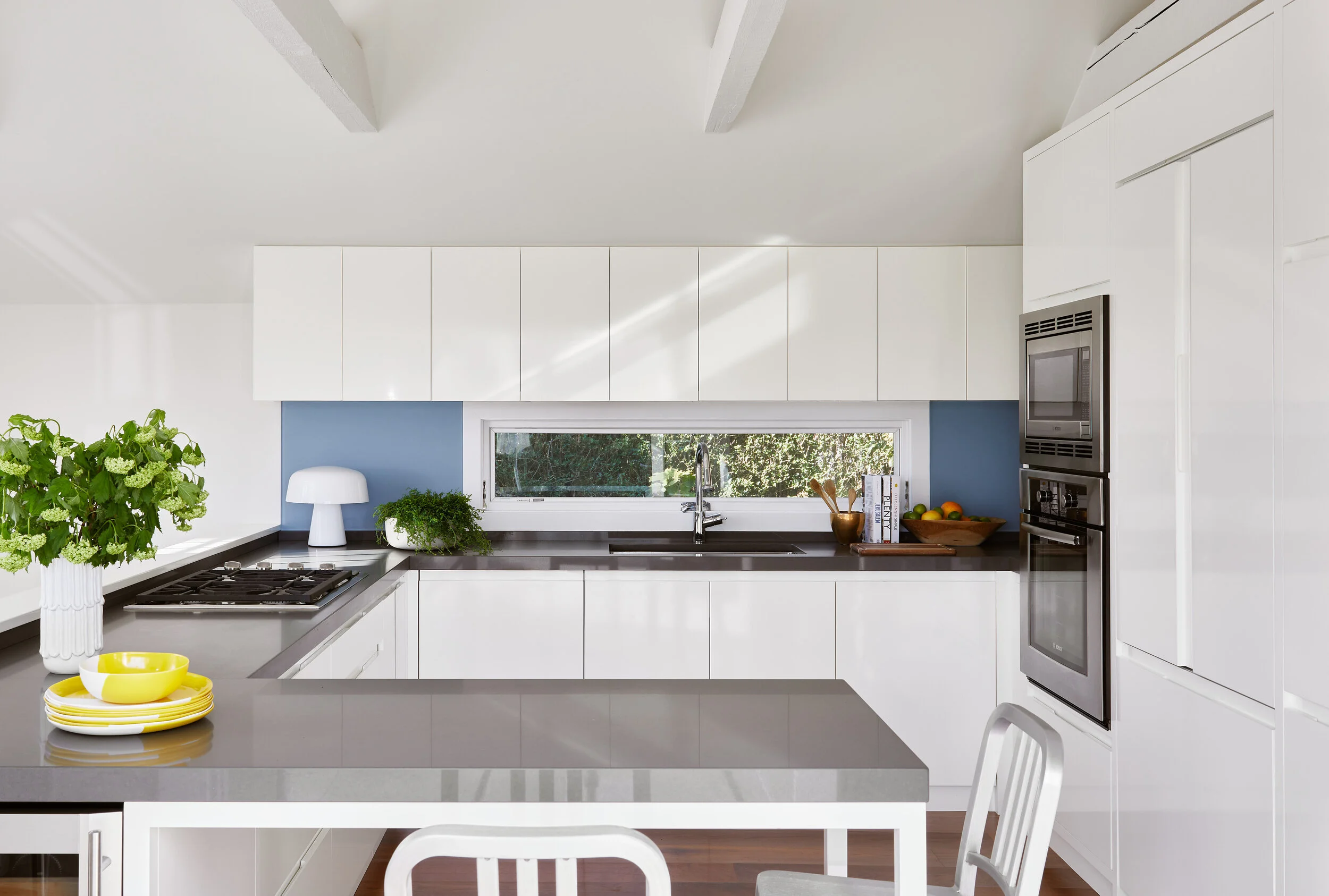 Modern white kitchen with black countertops, a window behind the sink, and colorful dishes and plants on the counter.