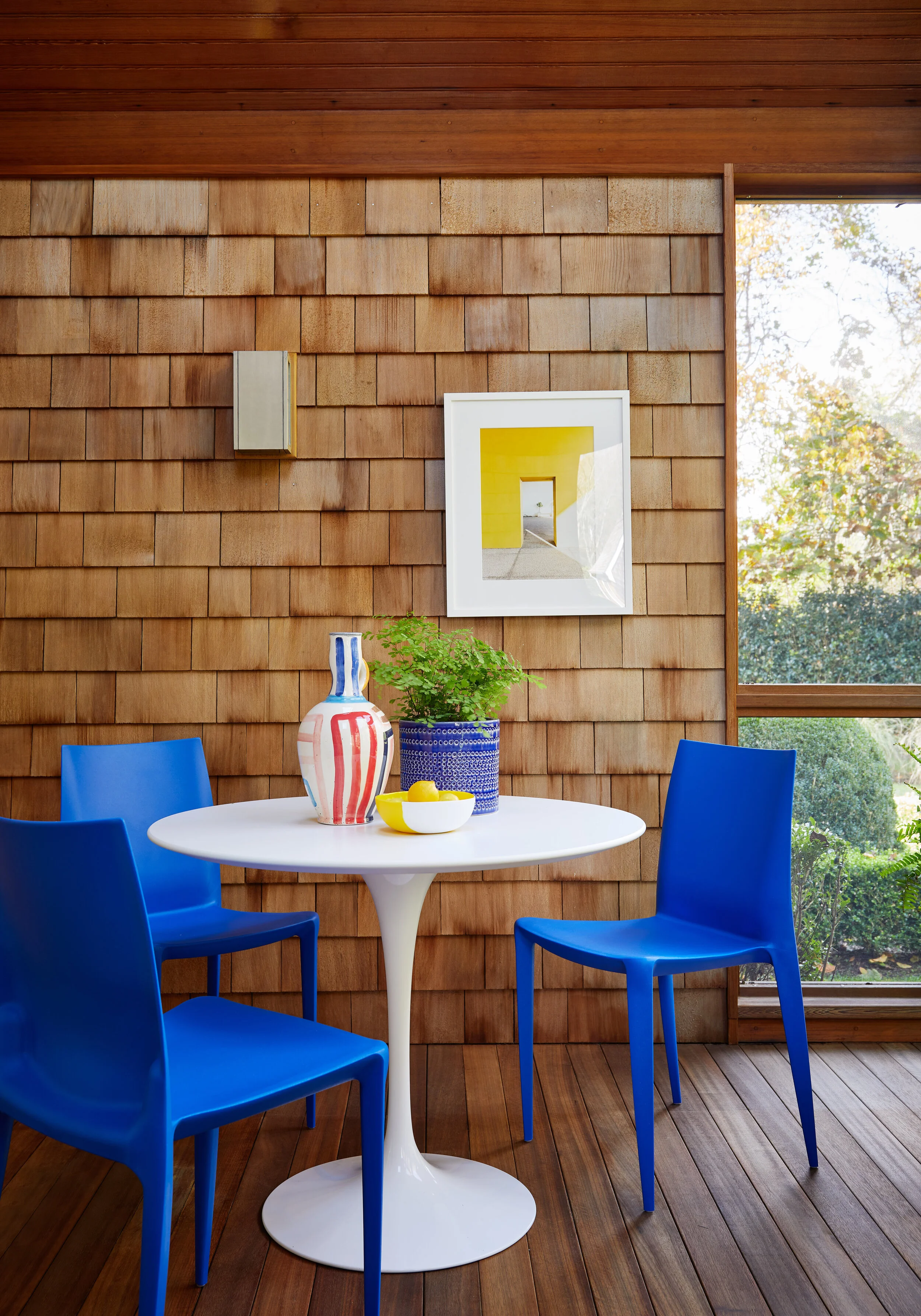 A cozy dining nook with a white round table, three bright blue chairs, a striped vase with a plant, a bowl with lemons, and a framed abstract artwork on a wooden shingle wall, with a large window showing greenery outside.