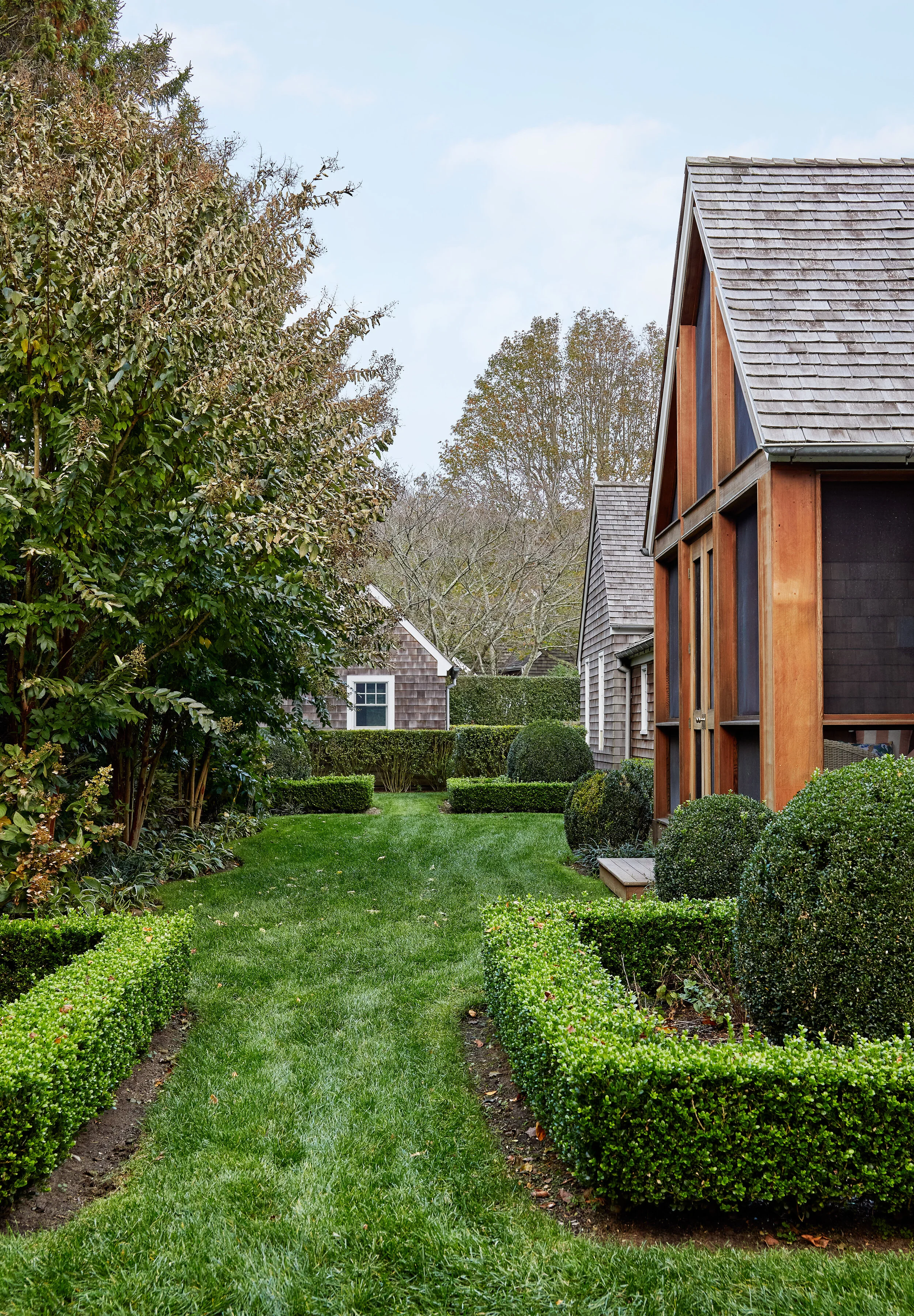 A neatly landscaped backyard with a narrow grass path, well-trimmed bushes, trees, and two houses with wooden and shingle exteriors under a partly cloudy sky.