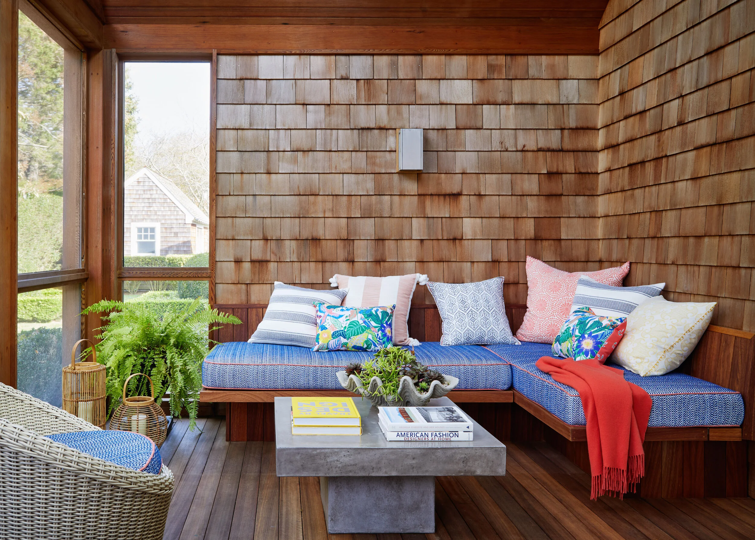 Sunroom with wooden walls, wicker chair, and a built-in wooden bench with cushions, pillows, and a red blanket, overlooking a garden and a neighboring house.