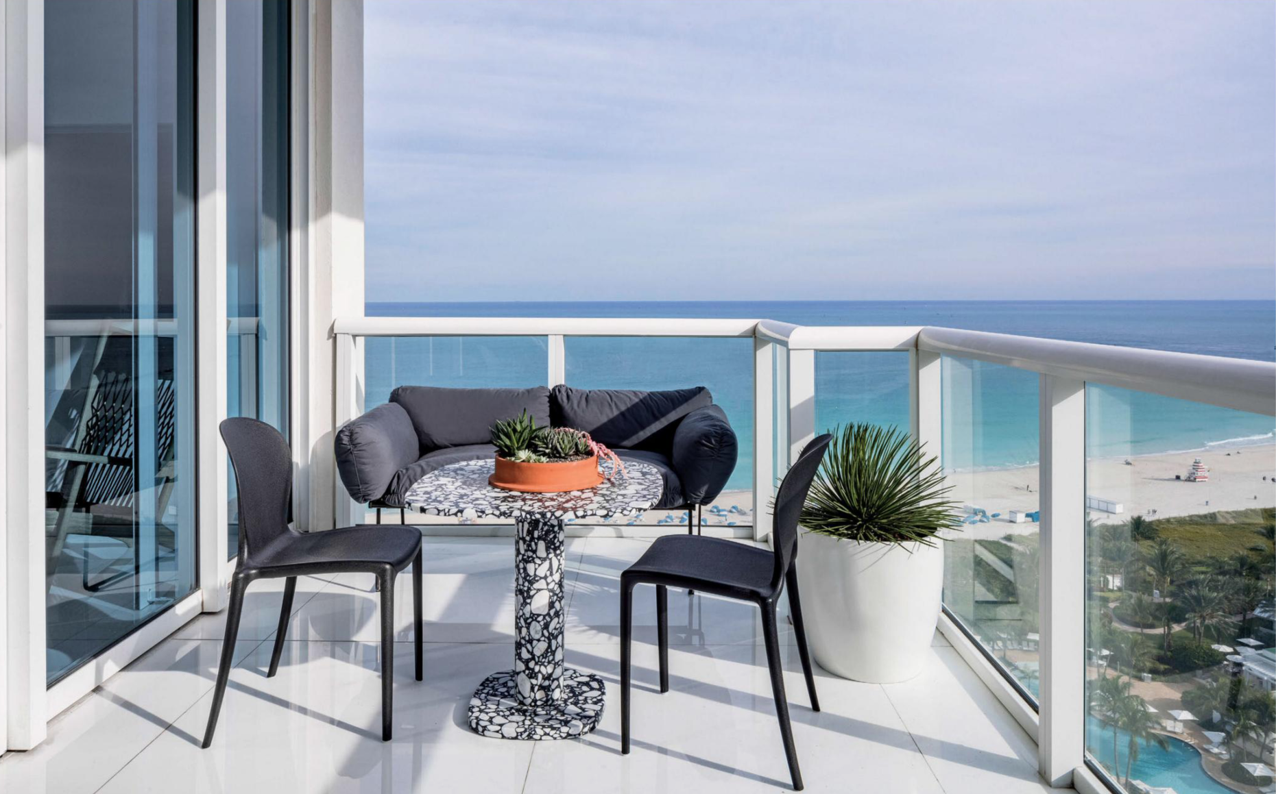 Balcony with two black chairs, a round table with a mosaic black and white top, a black sofa with gray cushions, and two potted plants overlooking a beach and ocean.