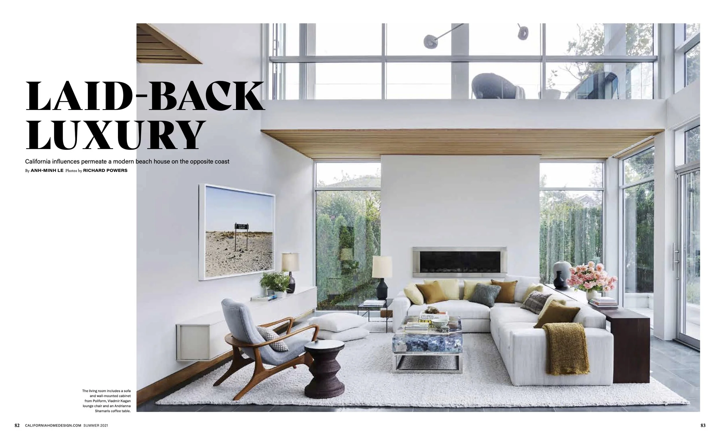 Bright modern living room with white walls and large windows, beige sectional sofa with colorful pillows, a gray armchair with wooden arms, a glass coffee table, side table with flowers, and a fireplace.