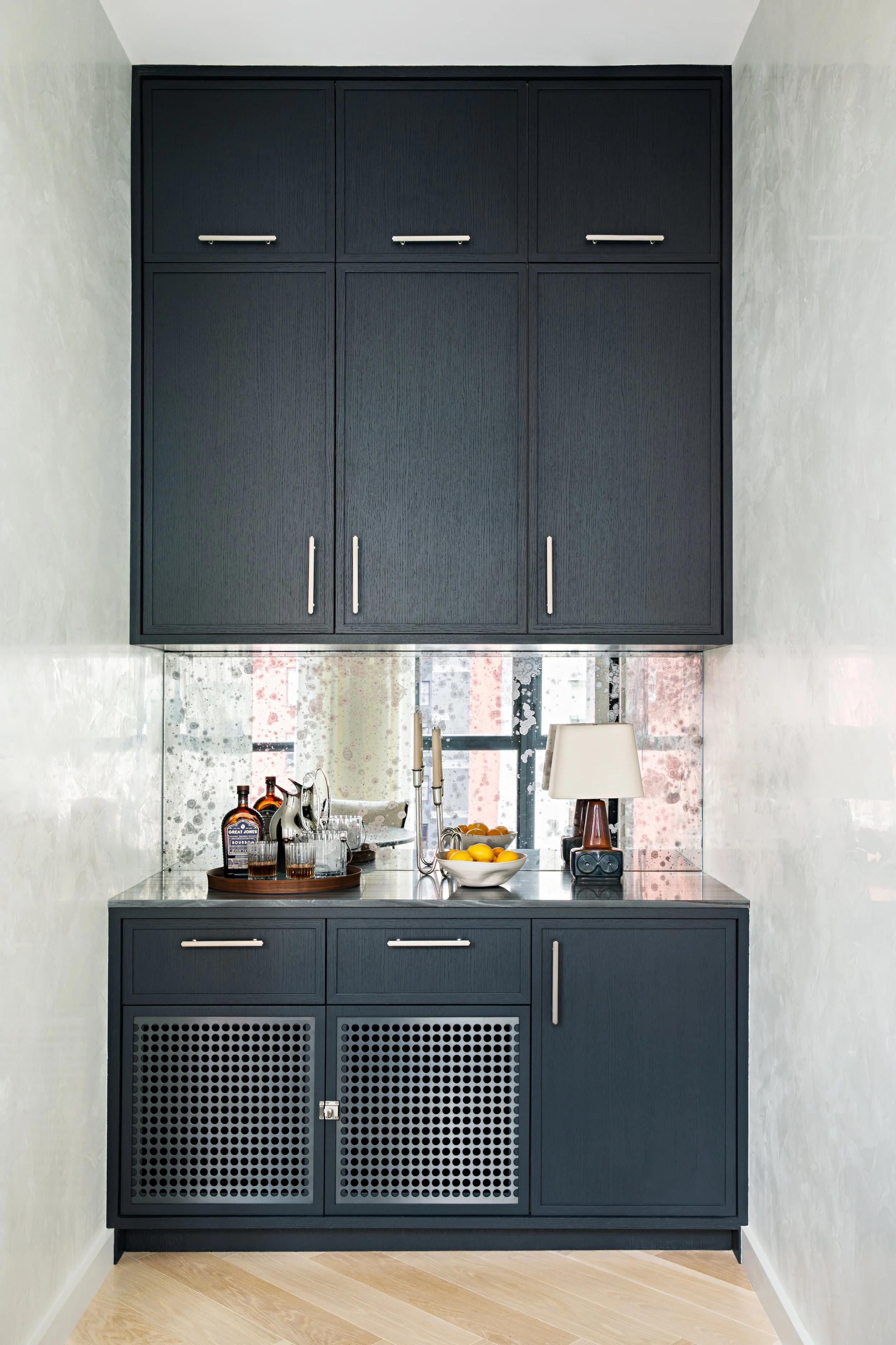 A built-in bar area with dark gray cabinets, a mirrored backsplash, a lamp, and a bowl of lemons on the countertop.