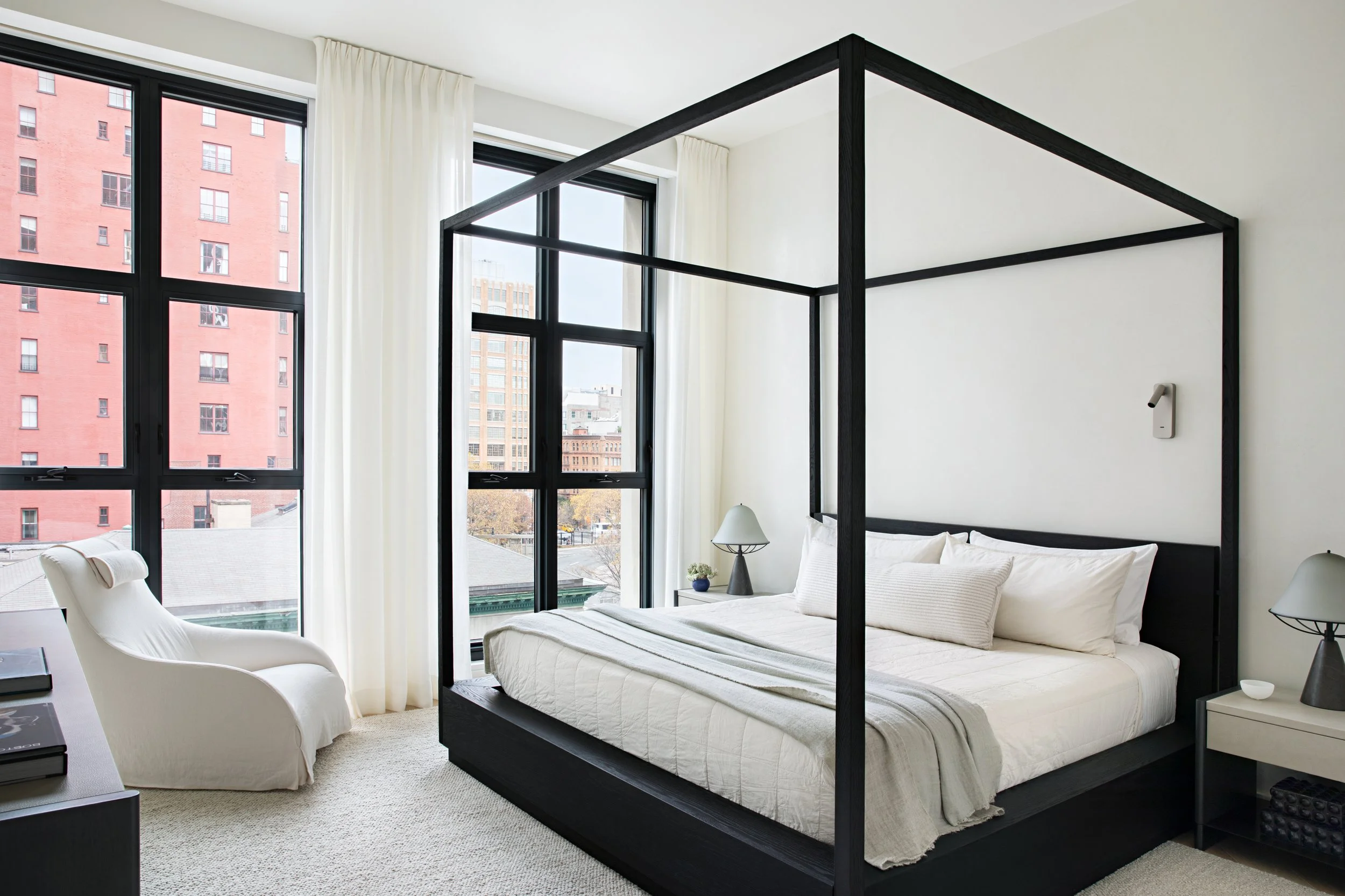 Modern bedroom with a black four-poster bed, white bedding, and large windows with city views. White curtains, a white armchair, and side tables with lamps.