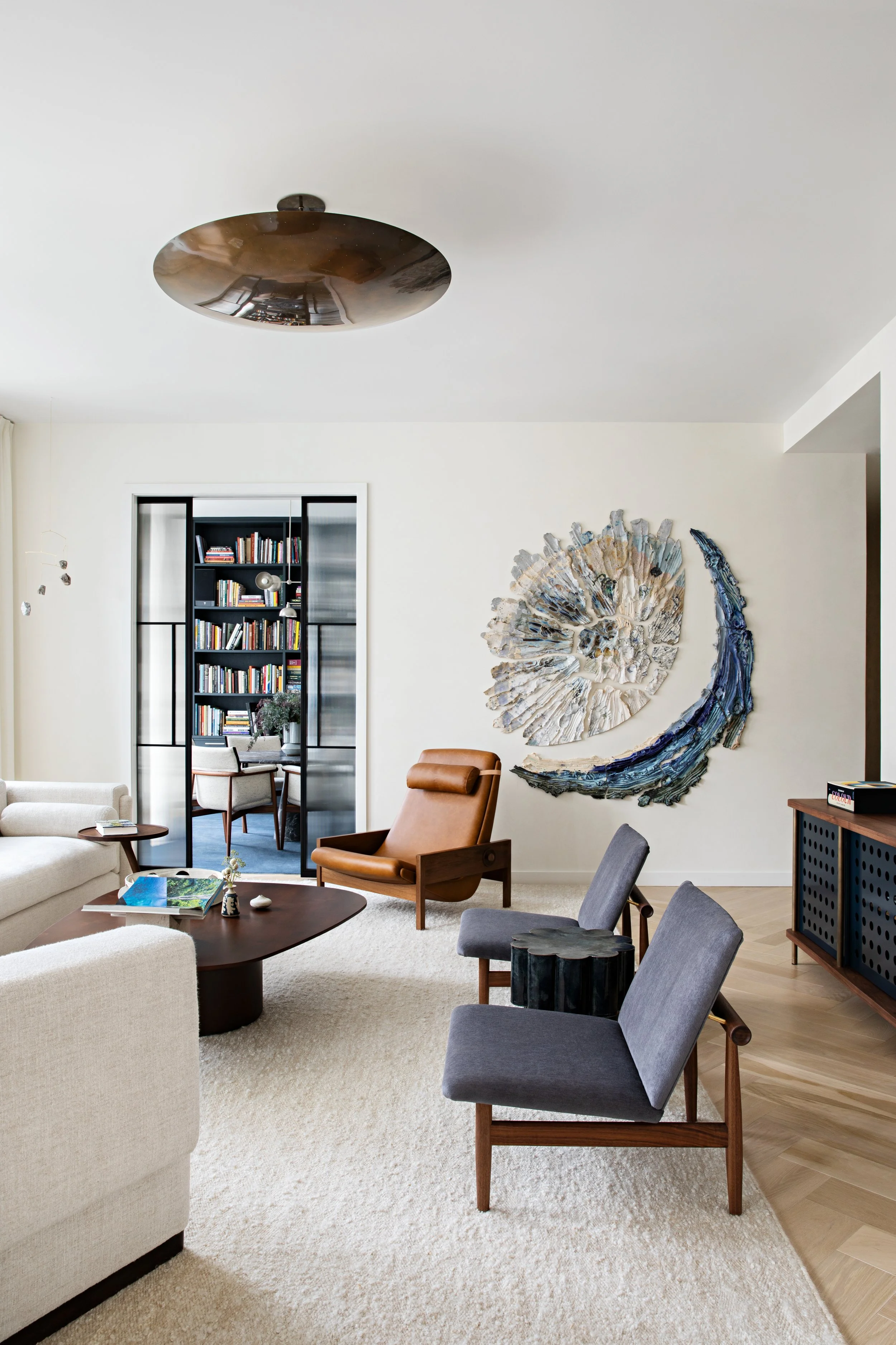 Modern living room with beige sofa, gray and brown chairs, a dark wood coffee table, a white rug, wall art resembling a shell and crescent moon, and a black bookshelf visible through a glass door.