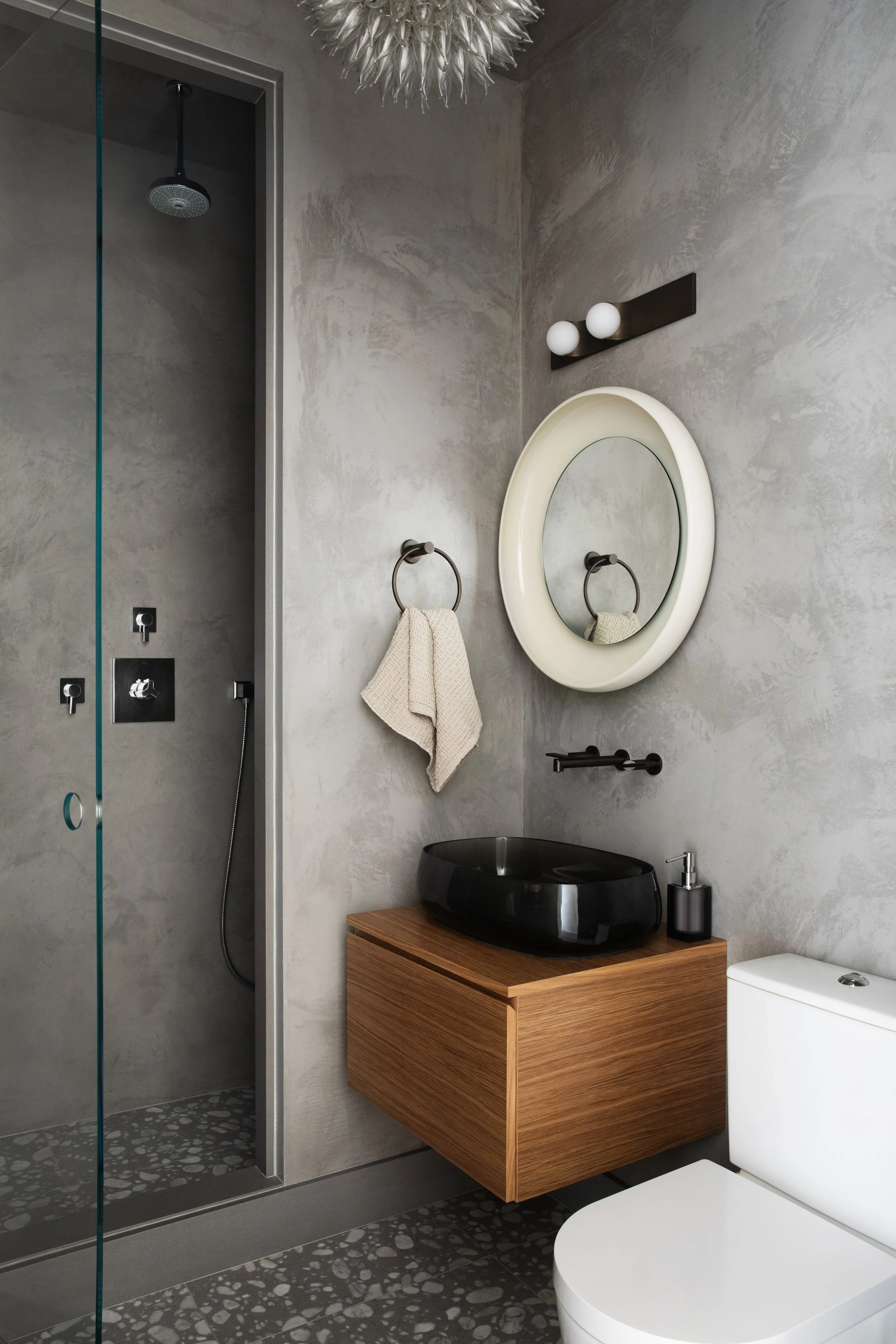 Modern bathroom with concrete textured walls, a glass shower with pebble floor, floating wooden vanity with black bowl sink, round mirror, towel ring with towel, wall light fixture, and white toilet.