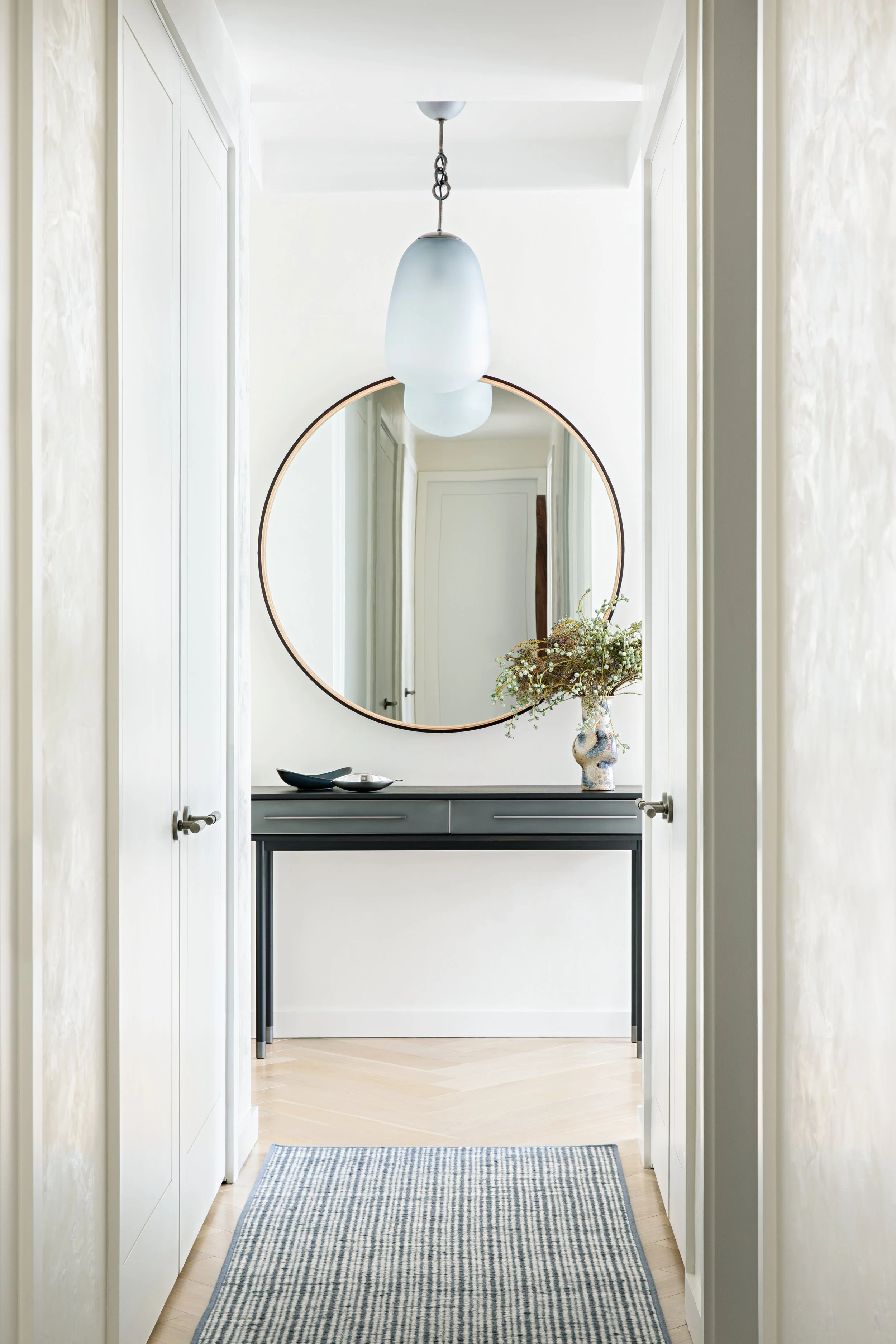 A hallway with a black console table, a circular mirror, a vase with flowers, a white hanging pendant light, and a blue striped rug on a light wood floor.