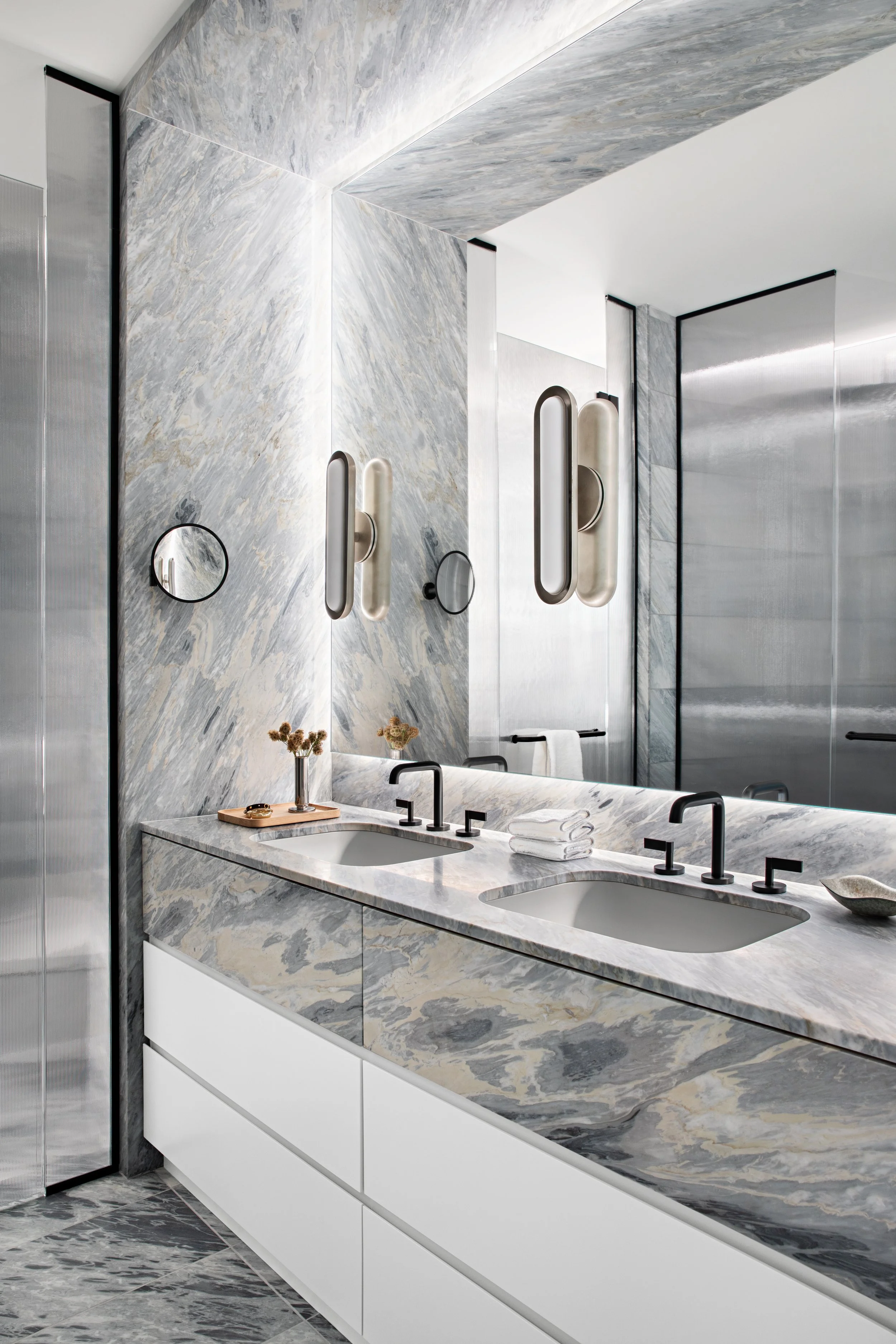 Modern bathroom with marble countertop and walls, double sinks, black faucets, large mirror, and decorative lighting fixtures.