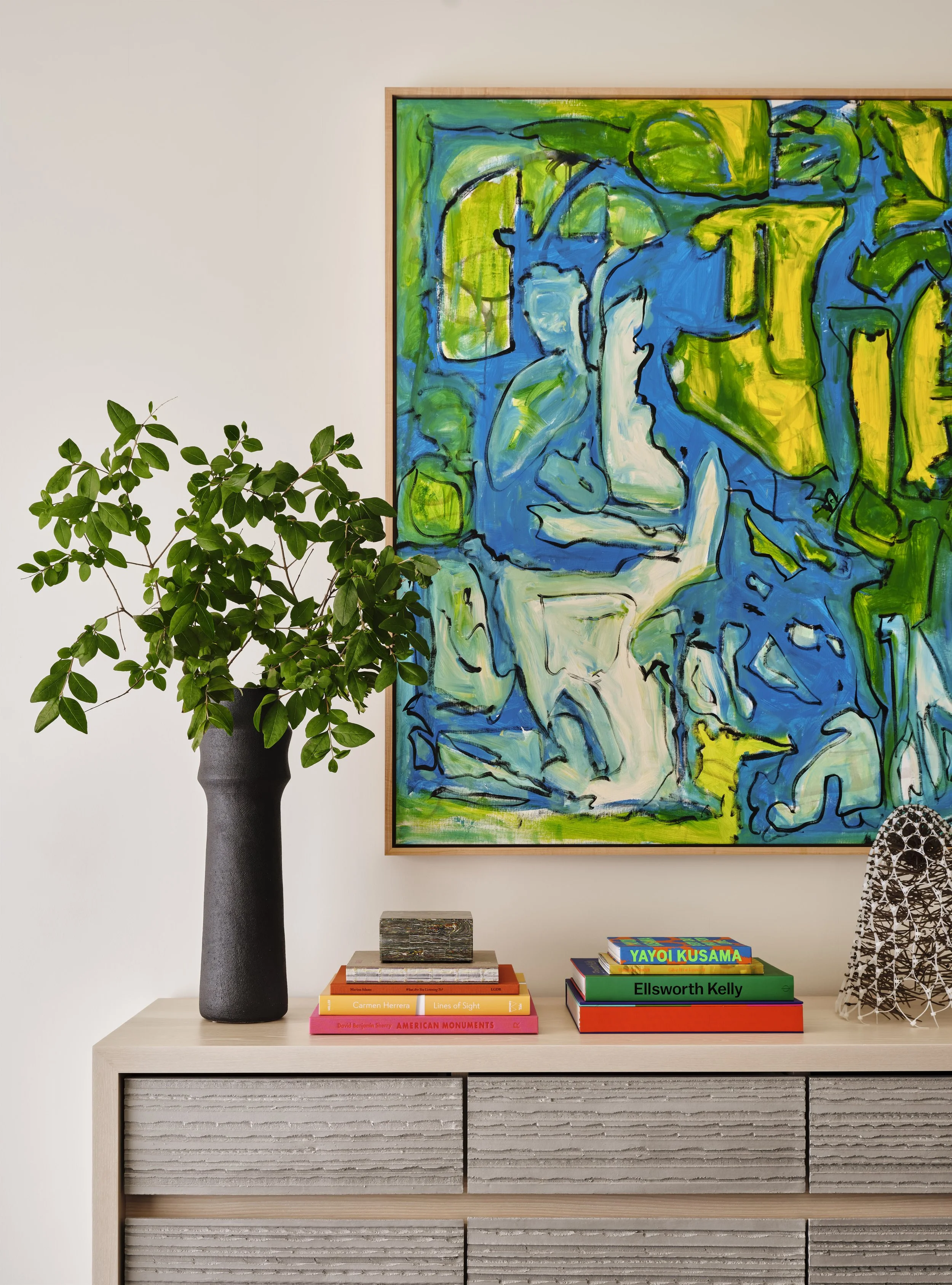 A modern sideboard with a large colorful abstract painting on the wall behind it. A tall black vase with a green leafy plant, and a stack of books including titles by Yayoi Kusama and Ellsworth Kelly, are on the sideboard.