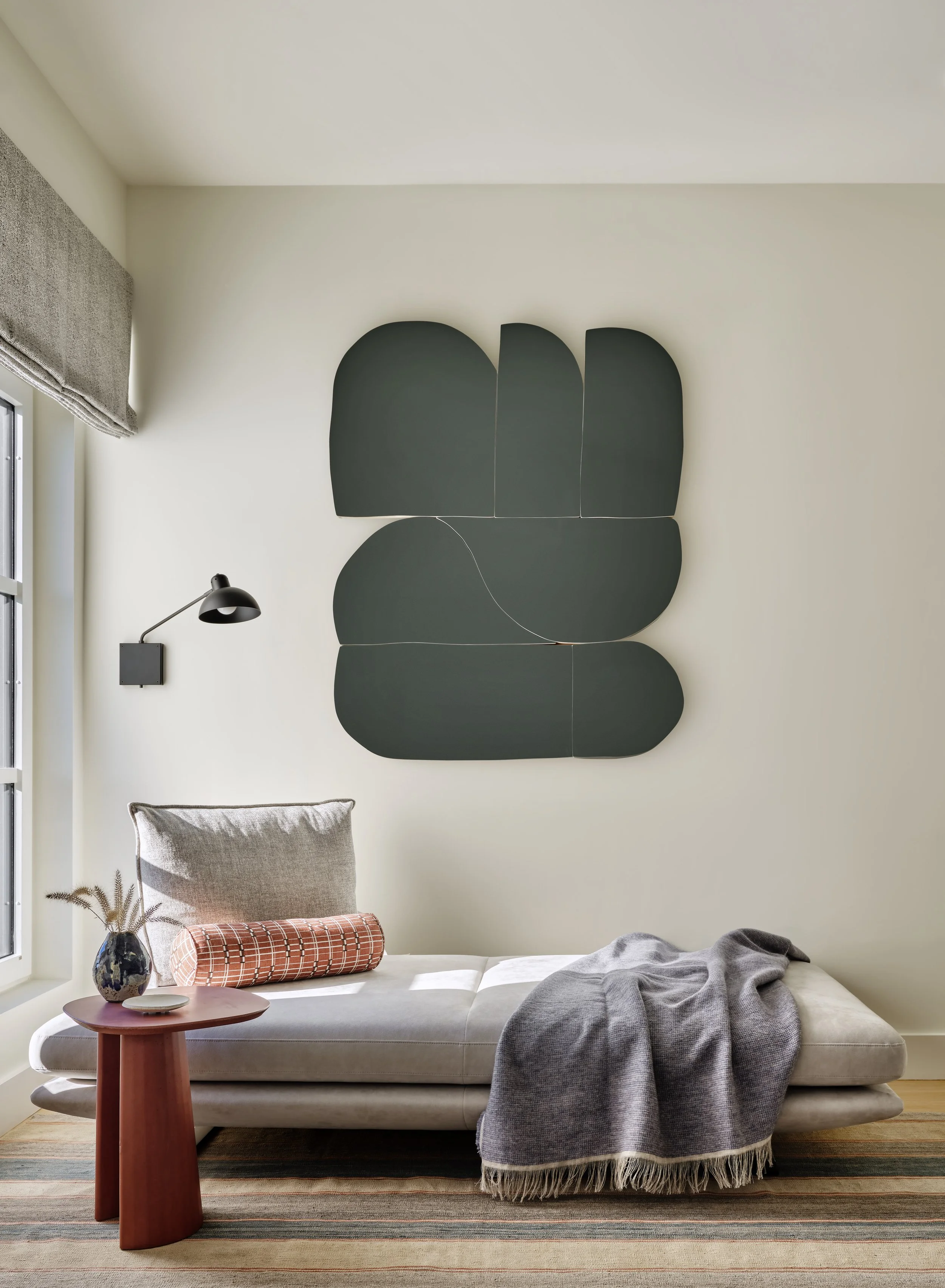 A modern living room corner with a beige chaise lounge, a gray throw blanket, a plaid cylindrical pillow, a small red side table with a vase of dried flowers, a black wall-mounted reading lamp, and abstract black wall art on a cream wall near a windo