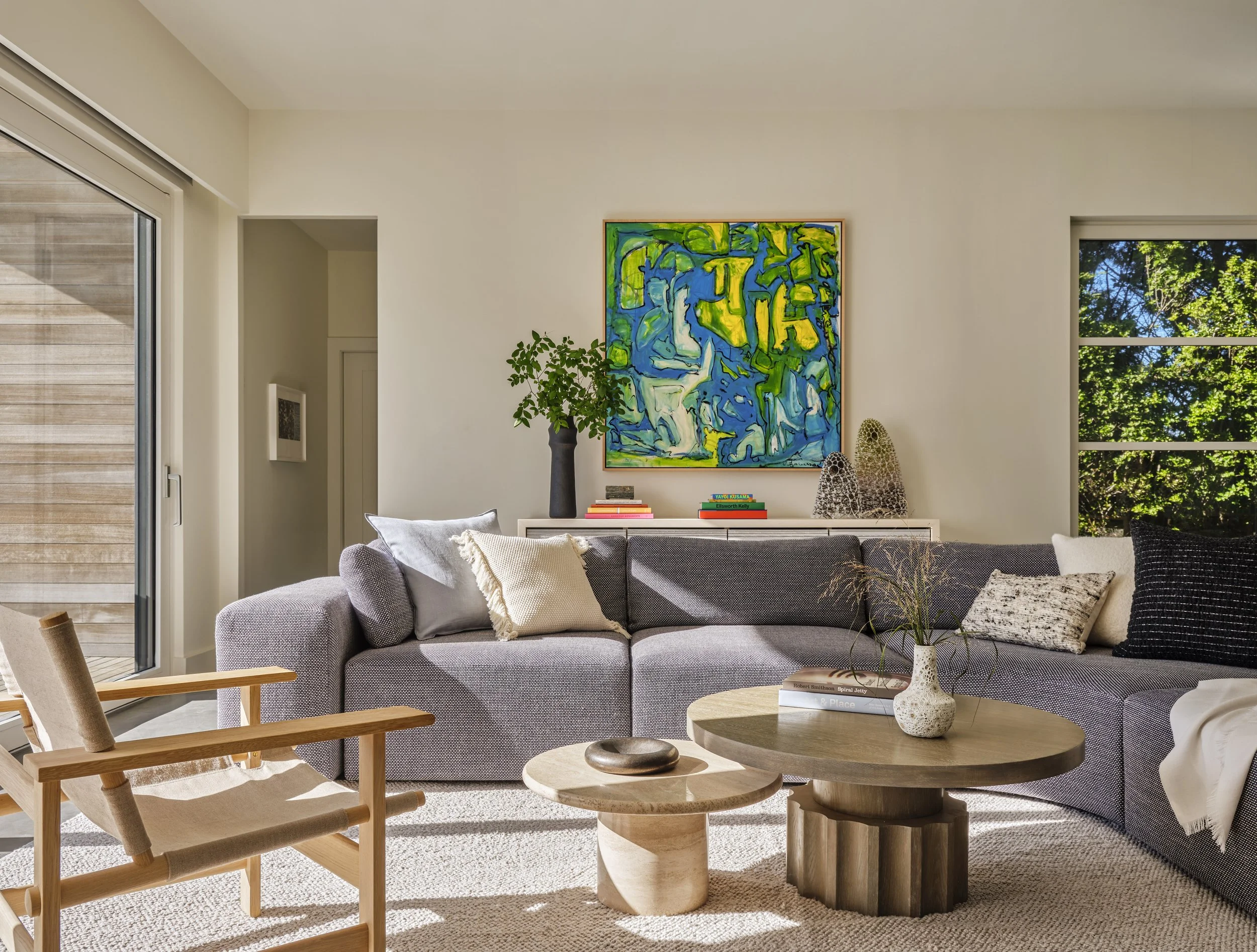 Living room with large gray sectional sofa, decorative pillows, wood coffee tables, abstract art painting, green plant, and windows showing greenery outside.