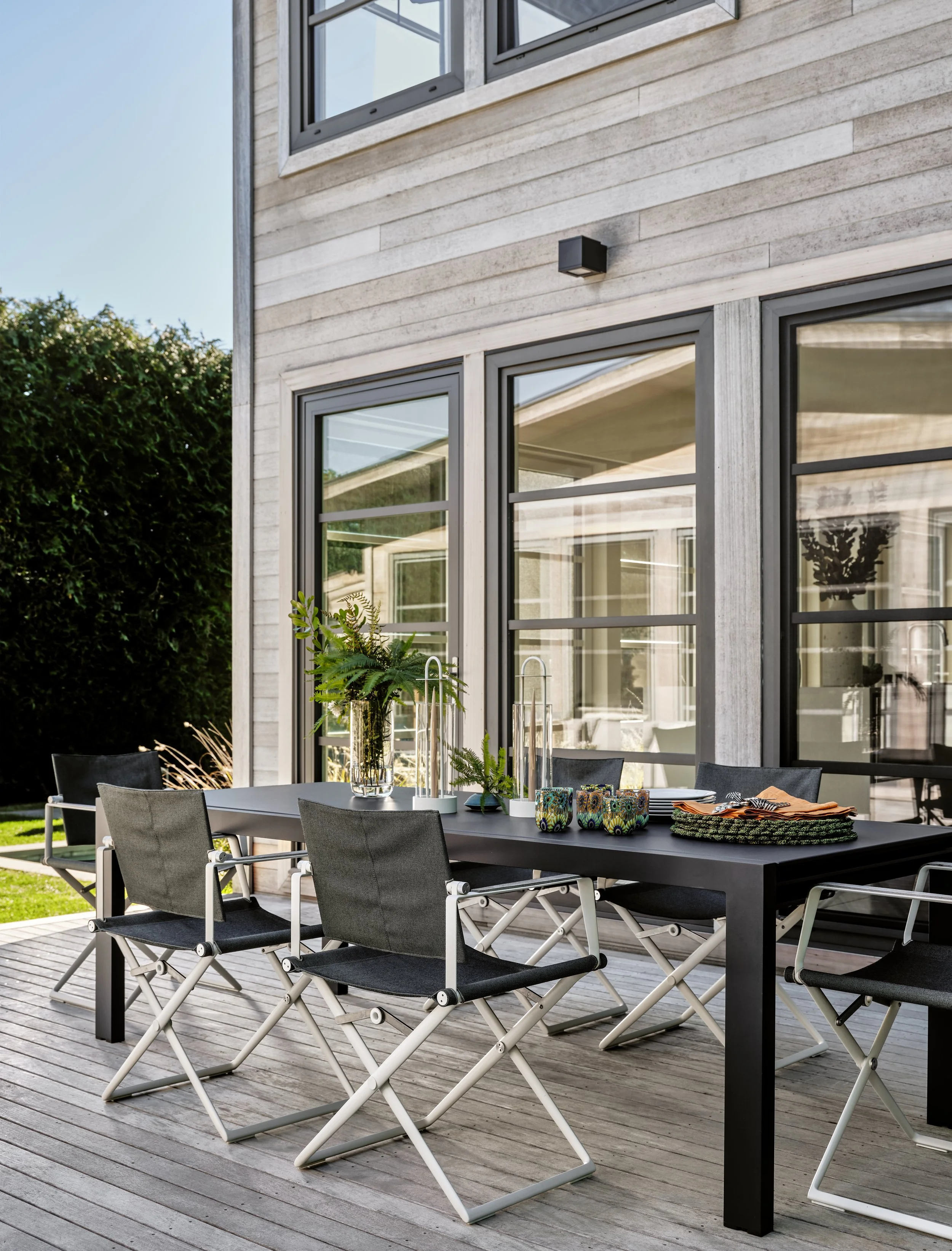 Modern outdoor patio dining area with black table, six black chairs, and decorative vases and plates, adjacent to a house with large windows and gray siding.