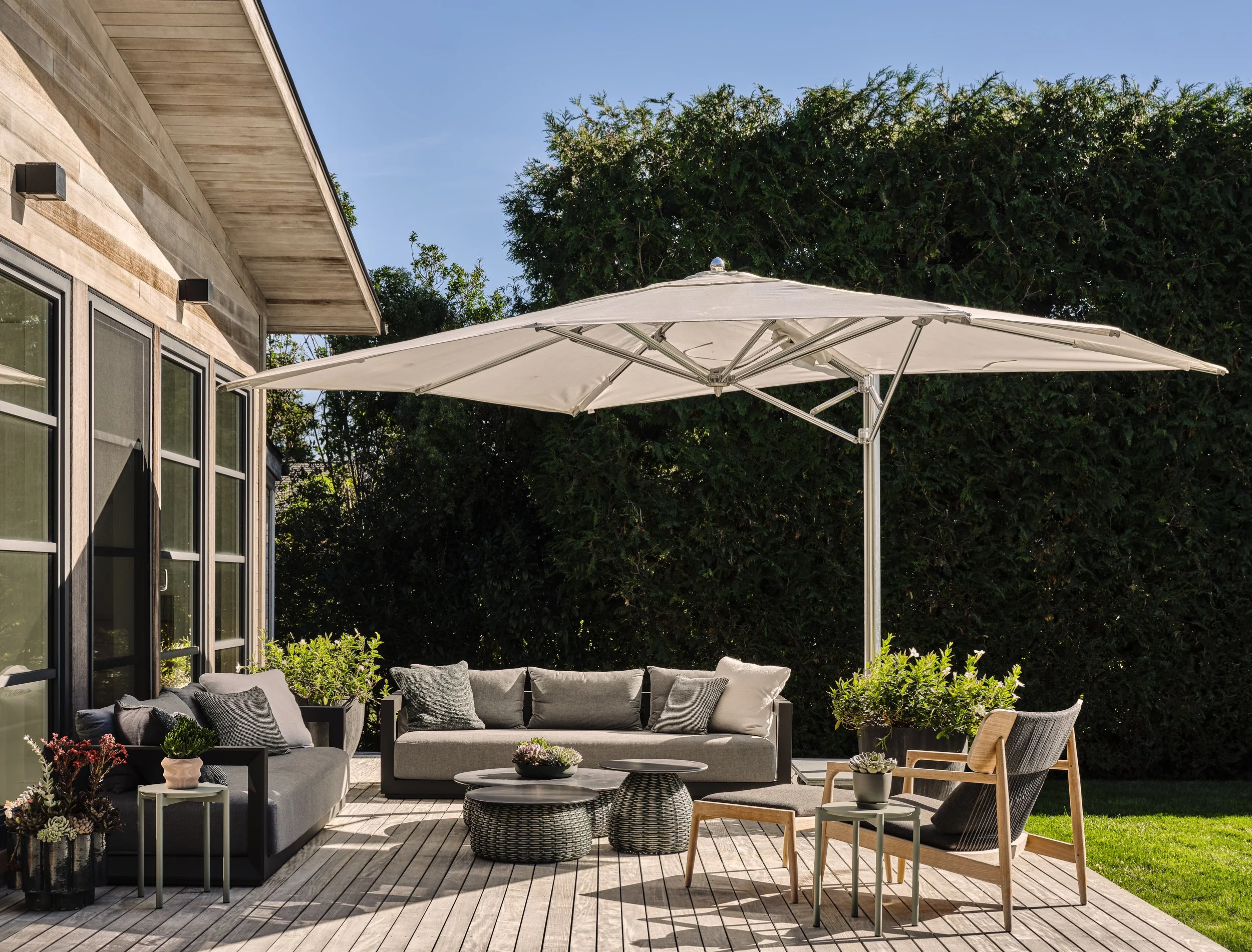 Outdoor patio with gray sofa, beige lounge chair, and coffee tables under a large white umbrella, surrounded by greenery.