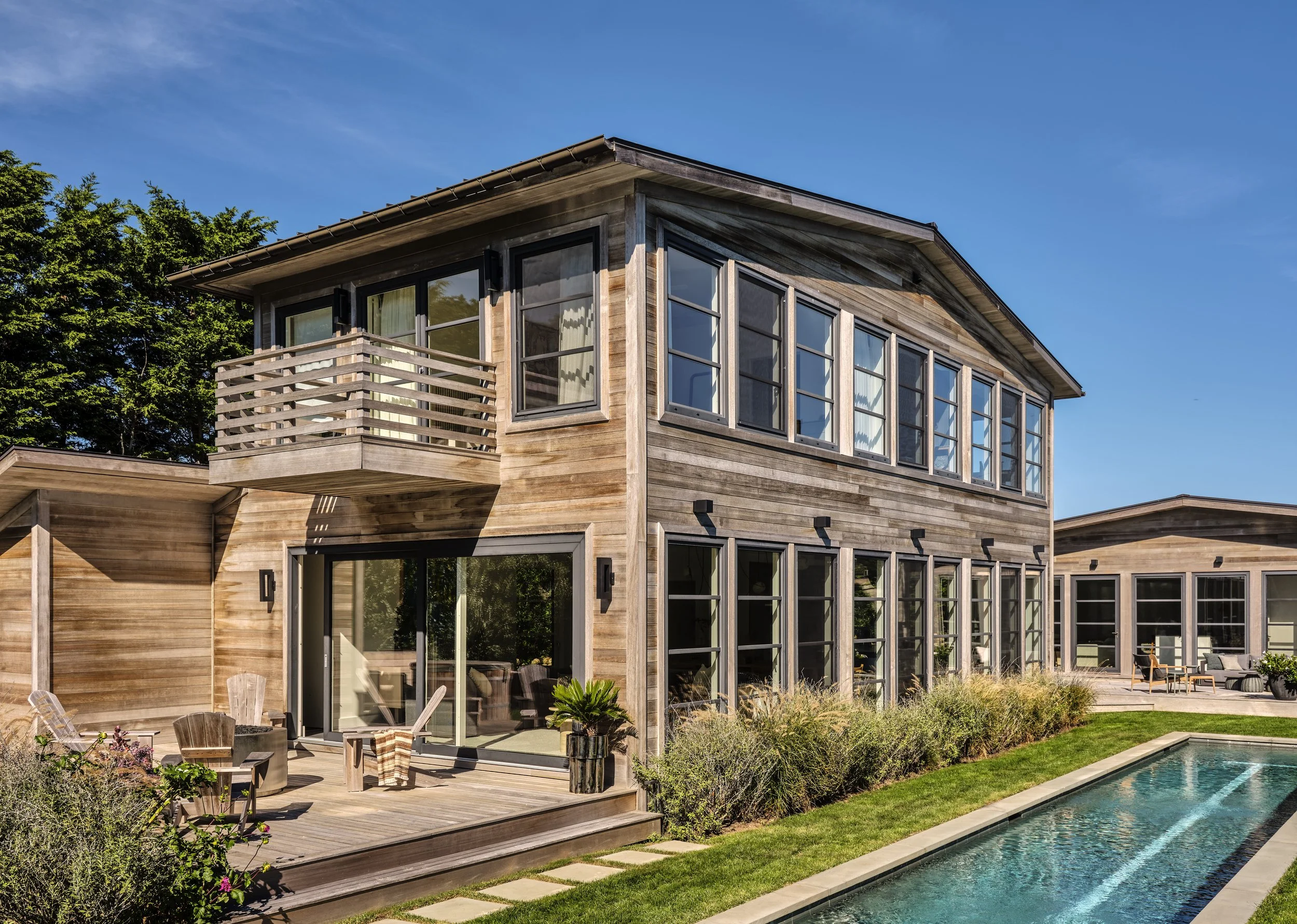 A modern, two-story house with a wood exterior, large windows, a balcony, and a backyard with a pool and outdoor seating.