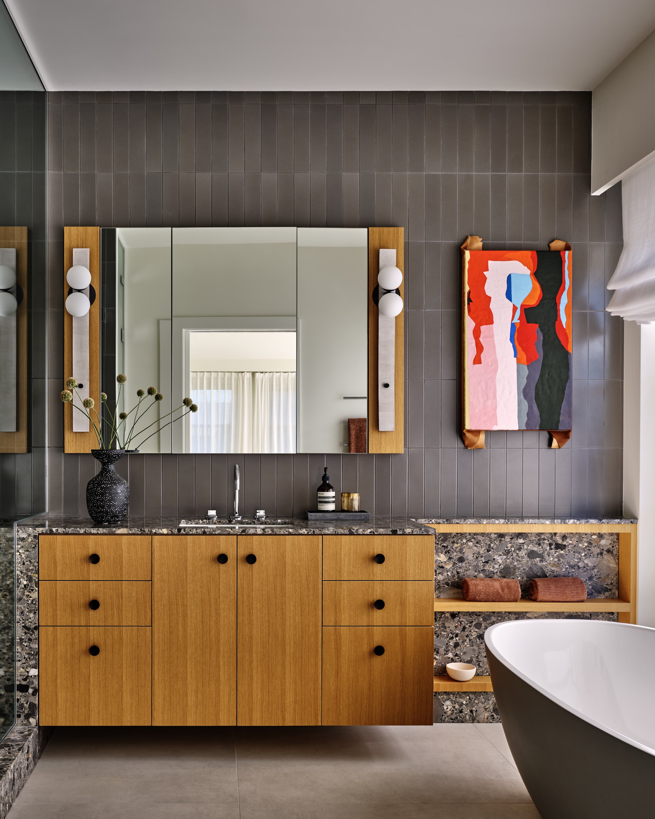 Modern bathroom with a wooden vanity, granite countertop, large mirror, abstract wall art, bathtub, and gray tiled wall.