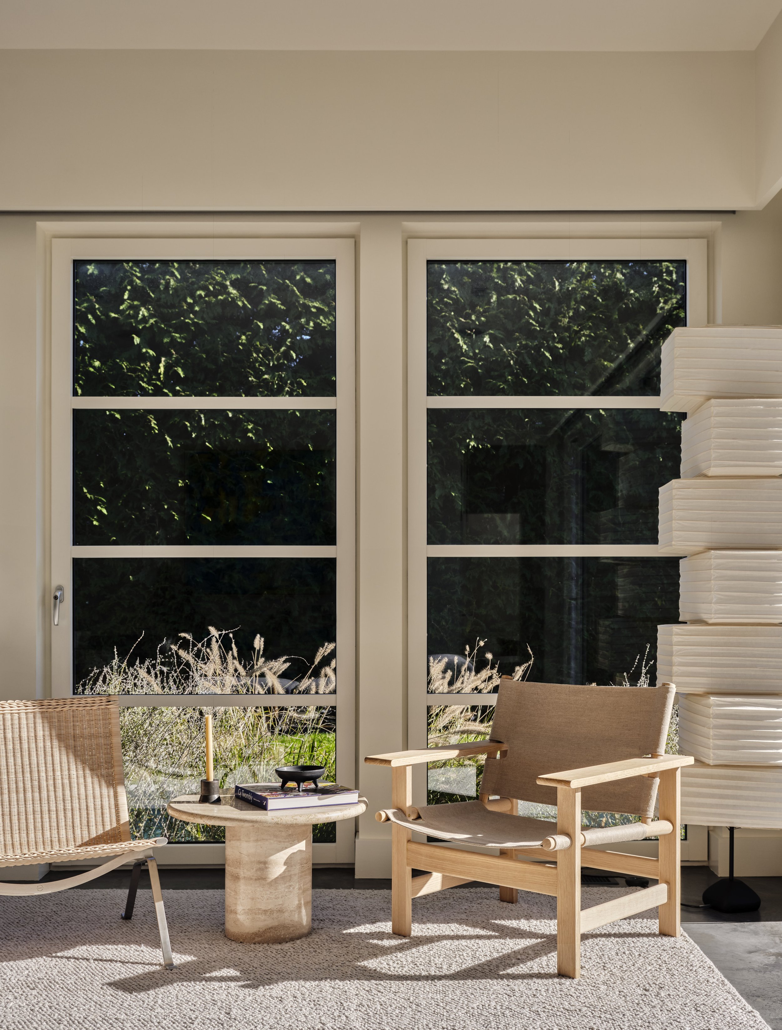 Living room with two large windows, a beige armchair, a wicker chair, a small round side table with a candle and books, and a stack of white foam boards on a black stand.