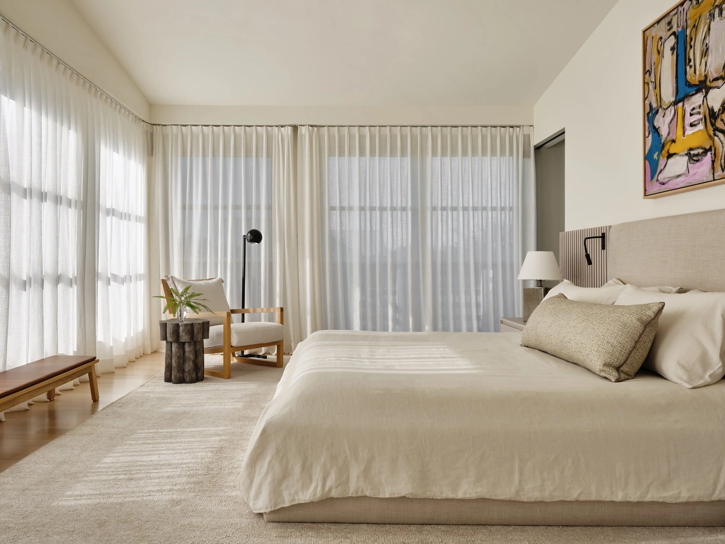 A modern, minimalist bedroom with a beige bed, a nightstand with a lamp, a chair with a throw pillow, a small side table with a plant, and large windows covered with sheer white curtains.