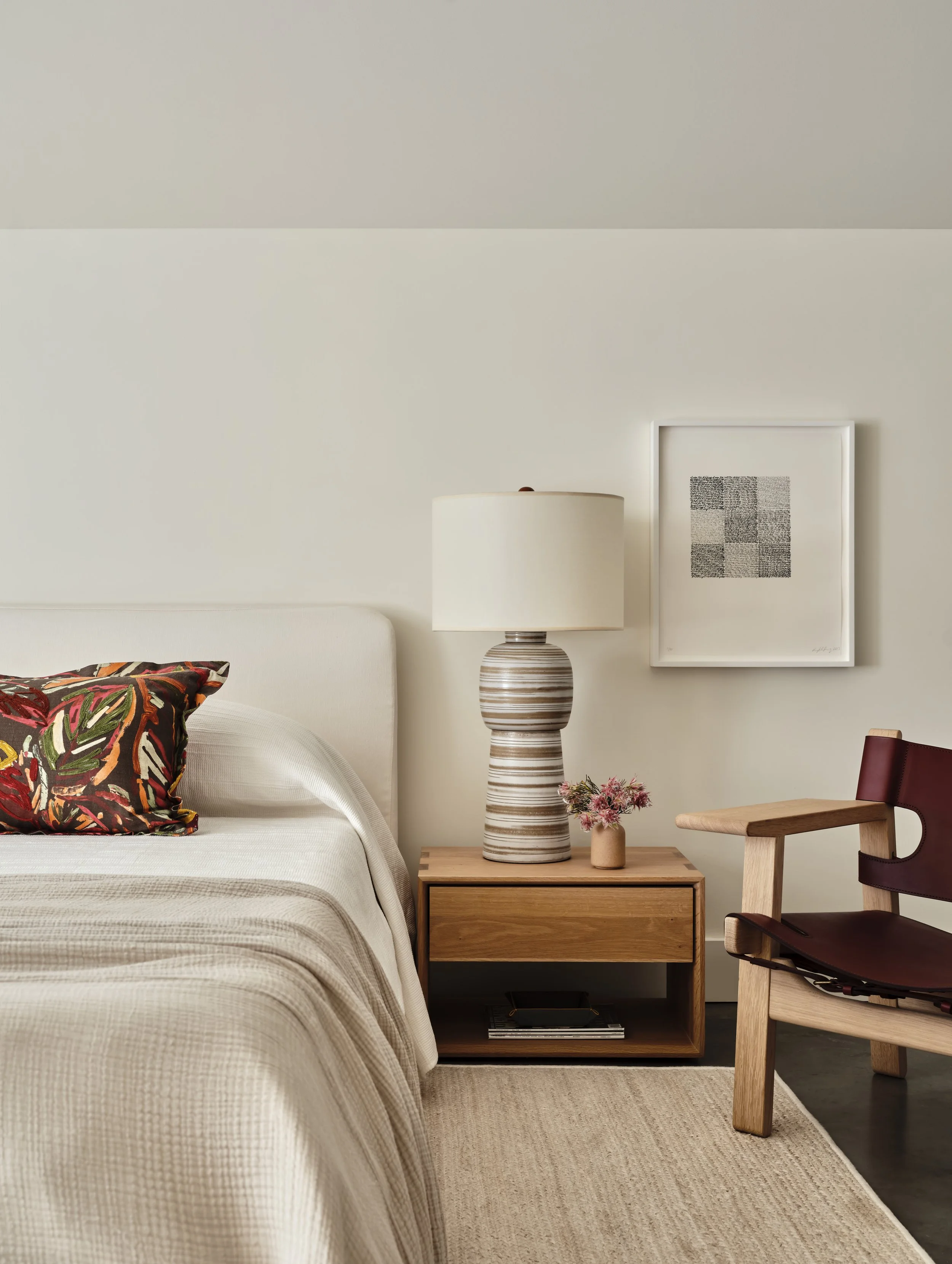 A bedroom with a beige bed, a colorful pillow, a wooden nightstand with a striped lamp, a small flower vase, and a framed black-and-white artwork on the wall. A wooden chair with a dark red leather seat is beside the nightstand.