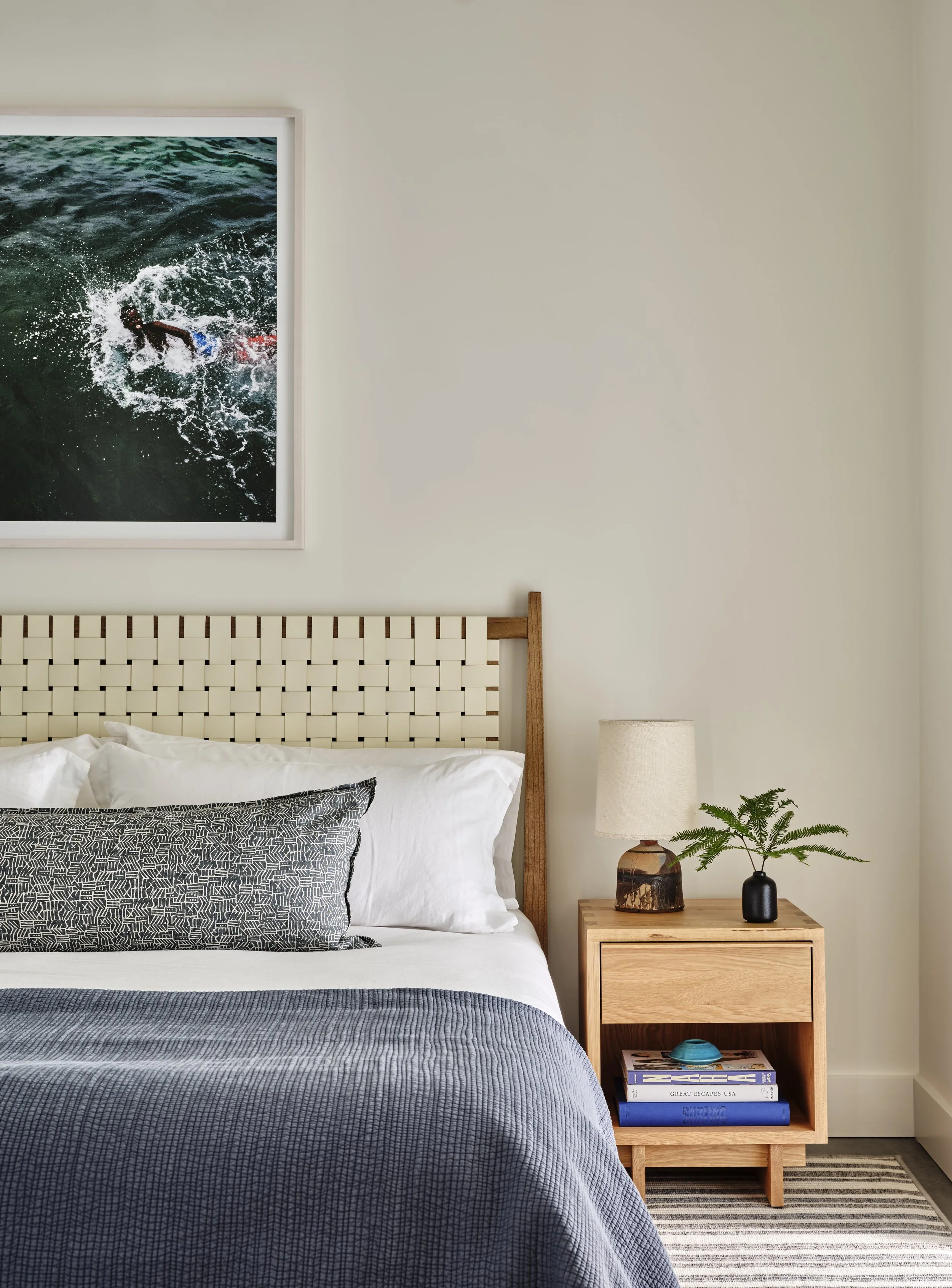 A bedroom with a wooden headboard, a bed with white sheets and decorative pillows, a nightstand with a lamp and a small plant, and a framed photograph of someone swimming in the ocean on the wall.