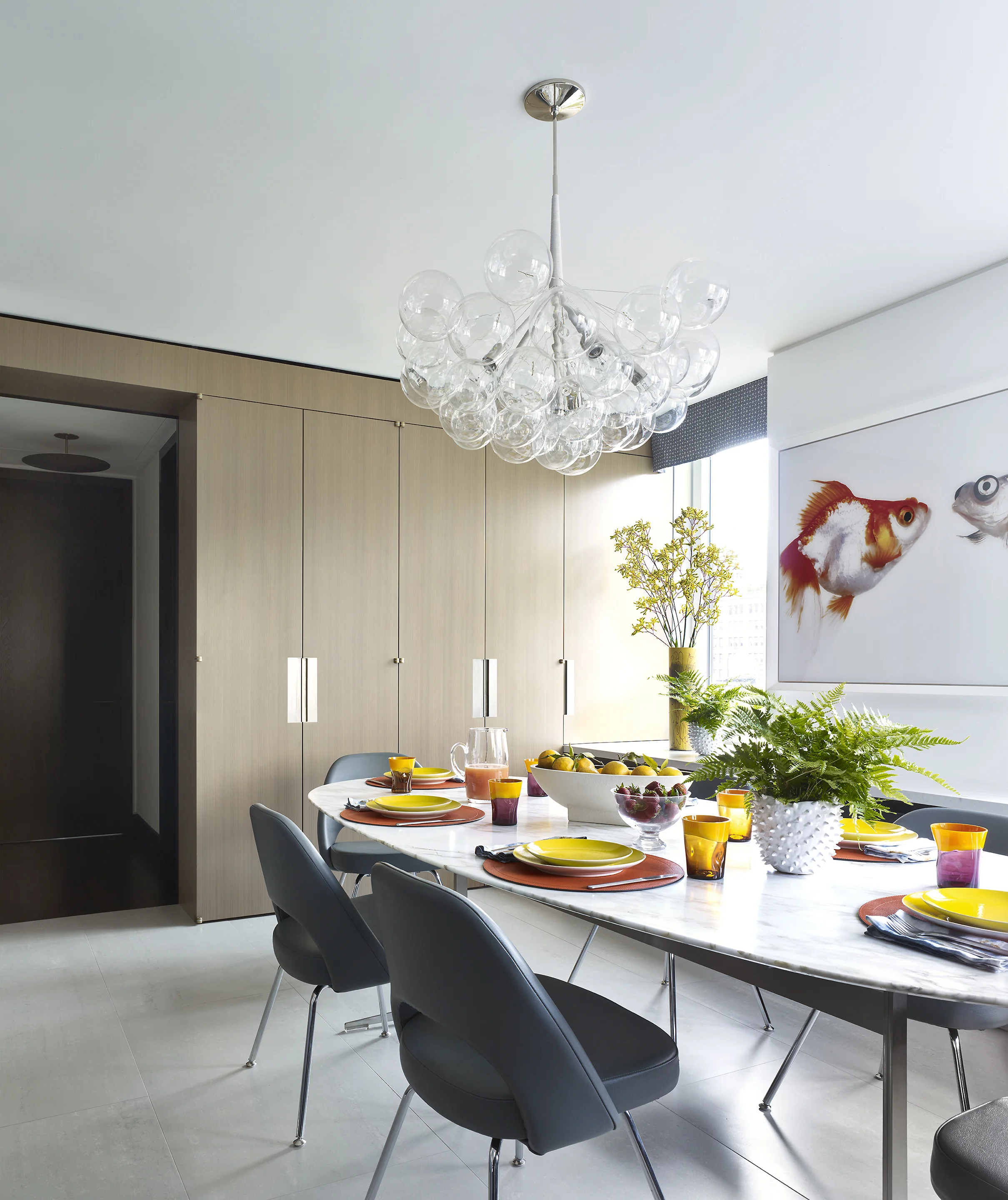Modern dining room with a white marble table set with yellow, orange, and purple dishes, surrounded by gray chairs, with a chandelier hanging from the ceiling, large windows, and wall art of colorful fish.