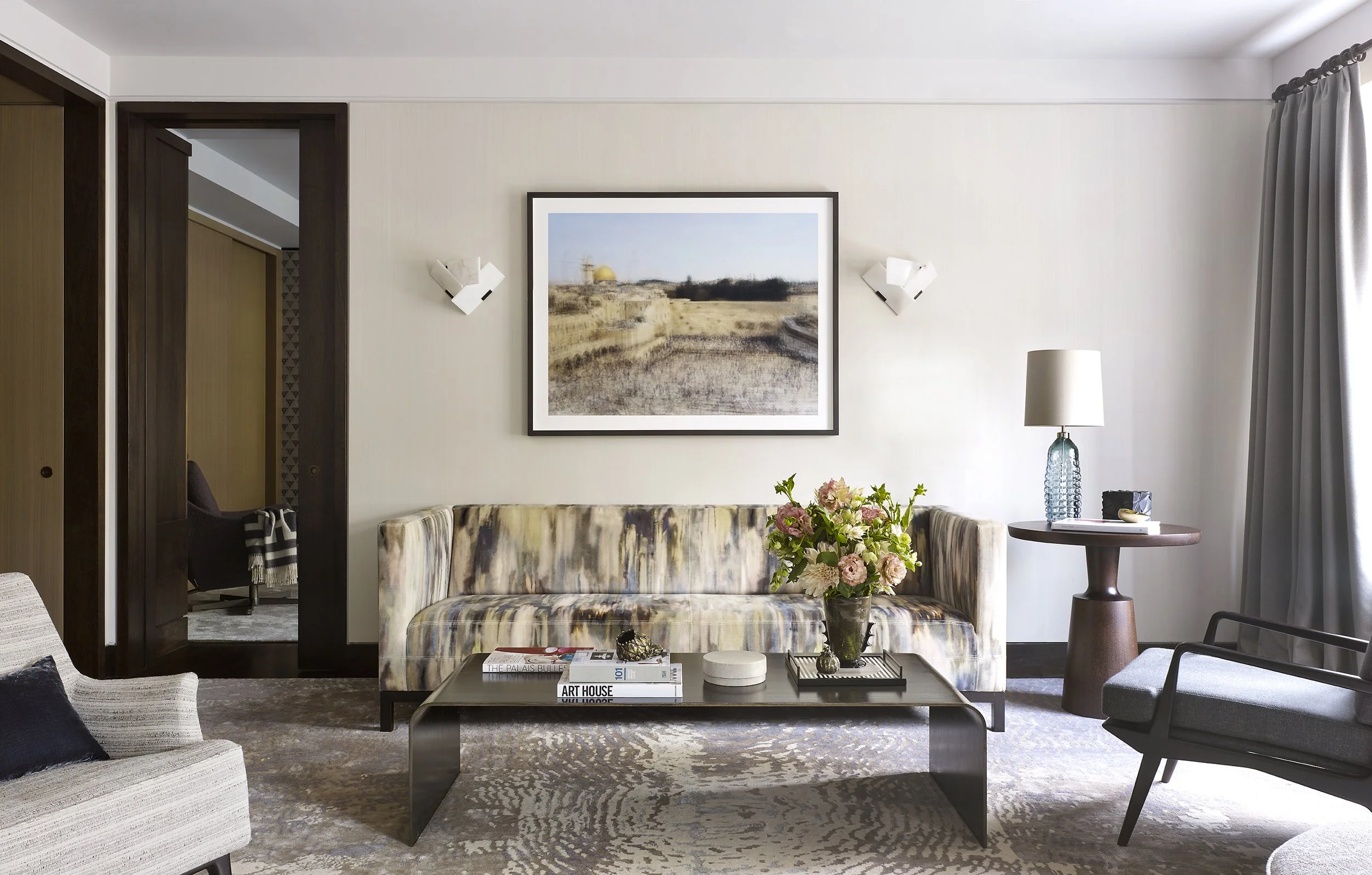 A living room with a patterned sofa, a coffee table with books and flowers, a side table with a lamp, and a gray armchair, with a window on the right and an abstract landscape painting on the wall.