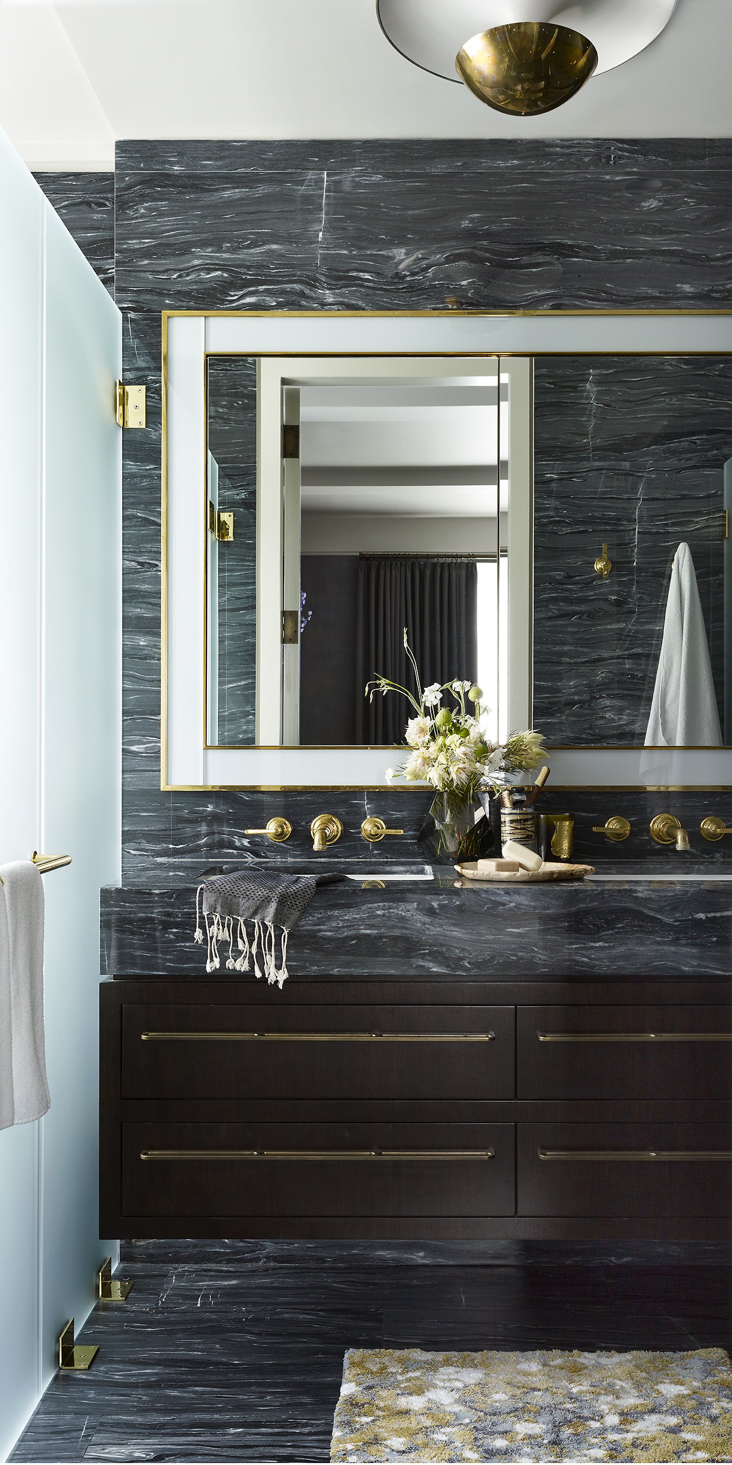 Modern bathroom with dark marble vanity, large mirror with gold trim, and gold fixtures. Decor includes a vase of white flowers and a small tray with toiletries. Visible towels hanging on gold hooks and a textured rug on the dark marble floor.