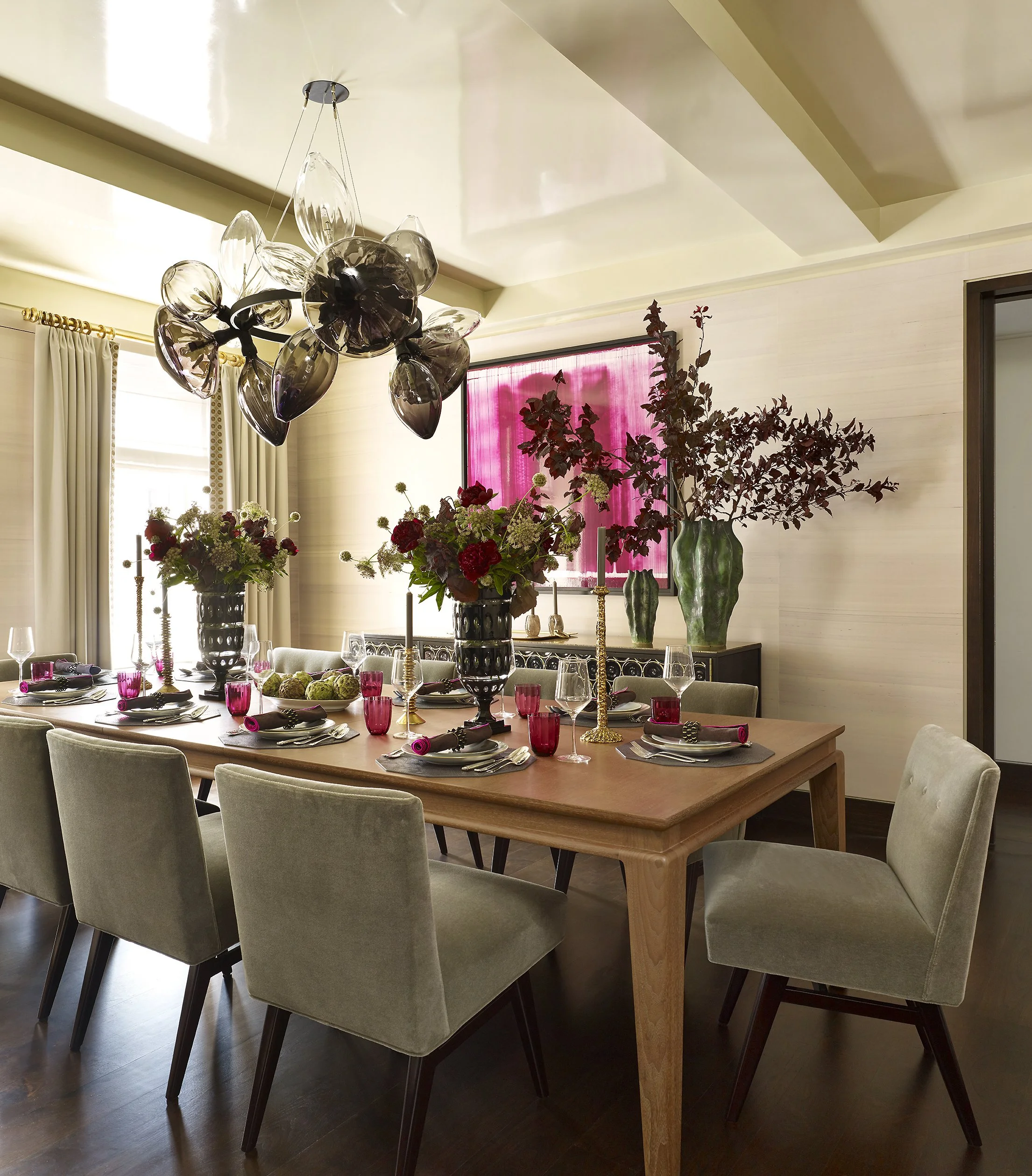 A dining room with a wooden table set with flowers, candles, glasses, and plates, and surrounded by beige upholstered chairs. There is a modern chandelier overhead, large windows with curtains, and decorative vases with dark red foliage and pink curt