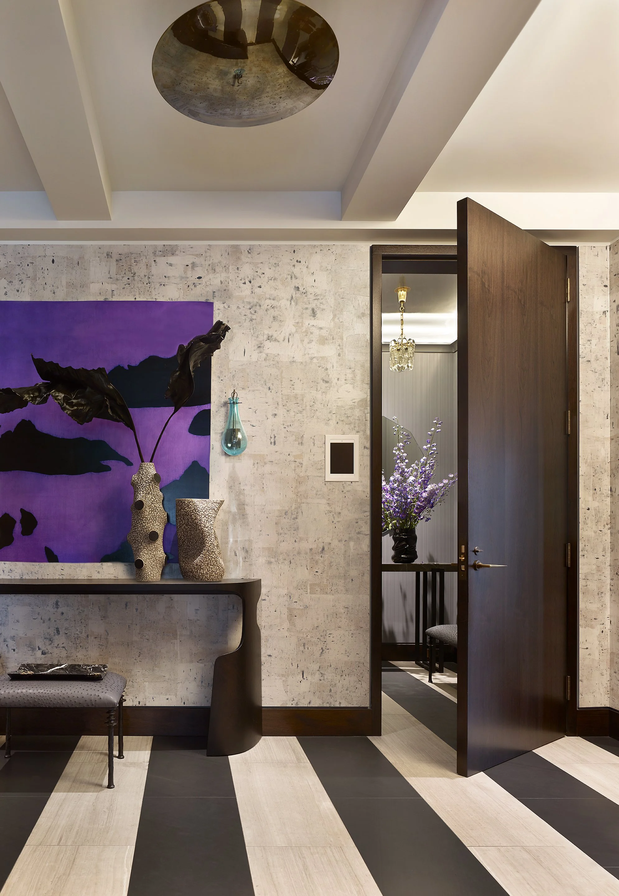 Interior view of a modern room with a textured wall, a console table with decorative vases, a purple abstract painting, and an open dark wood door leading to a room with a chandelier and a window with purple floral arrangements.