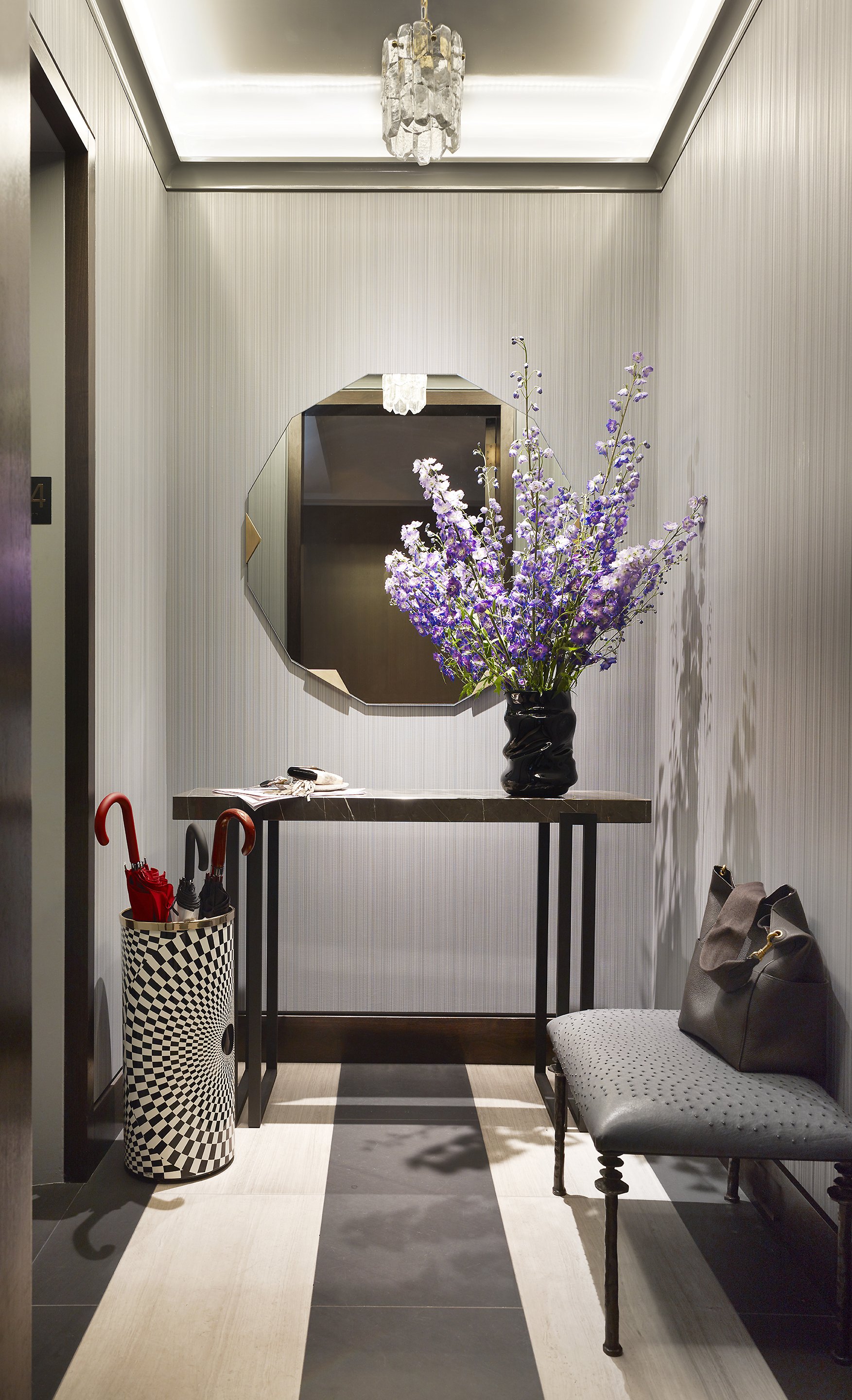 A modern hotel lobby corner with a black marble table decorated with a large vase of purple flowers, a spacious mirror on the wall, a bench with a gray handbag, and a black-and-white checkered umbrella stand with umbrellas, all under elegant lighting