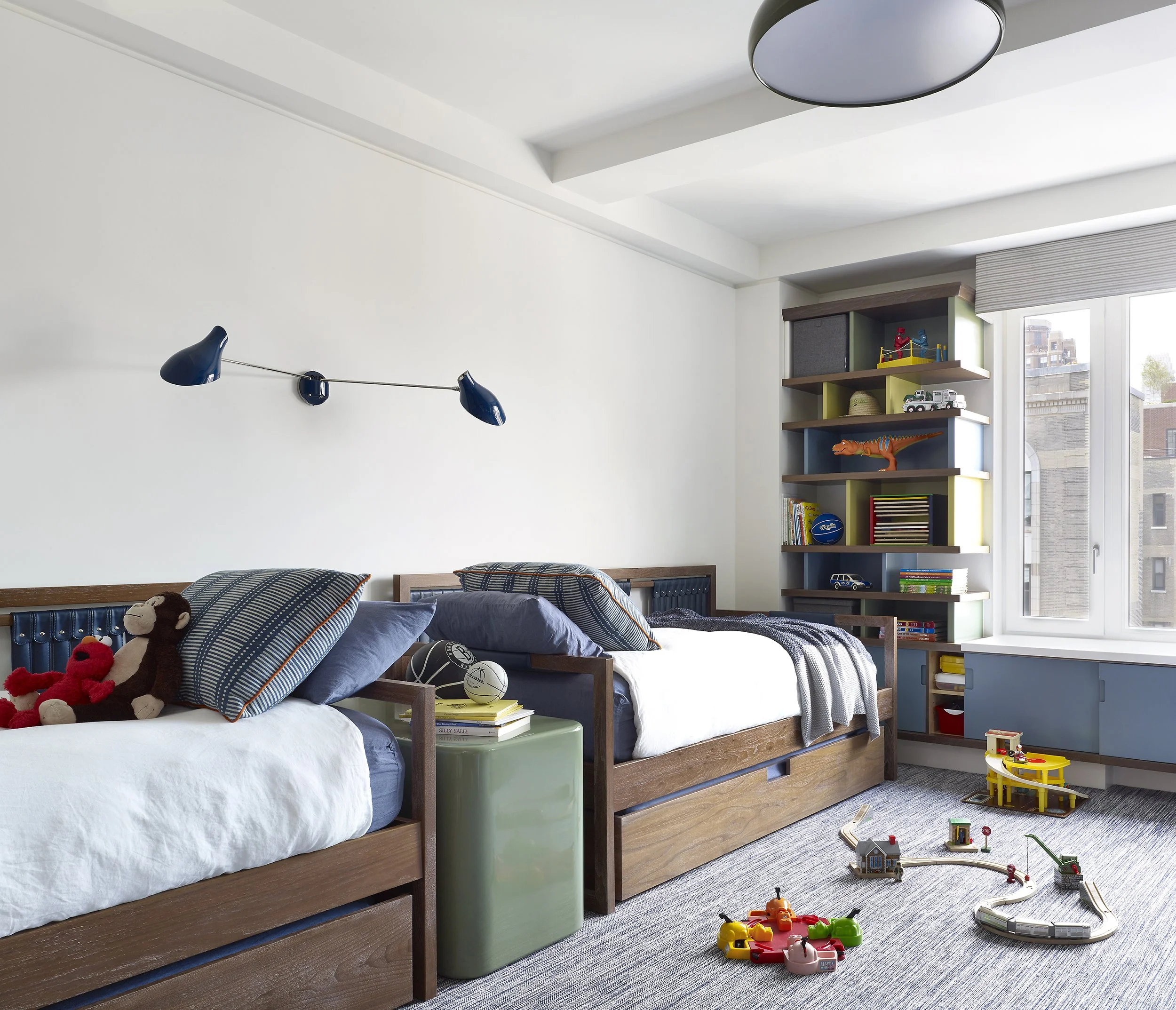 A children's bedroom with two beds, stuffed animals, toys, books, a green nightstand, a window with city view, and a colorful shelving unit filled with toys and books.