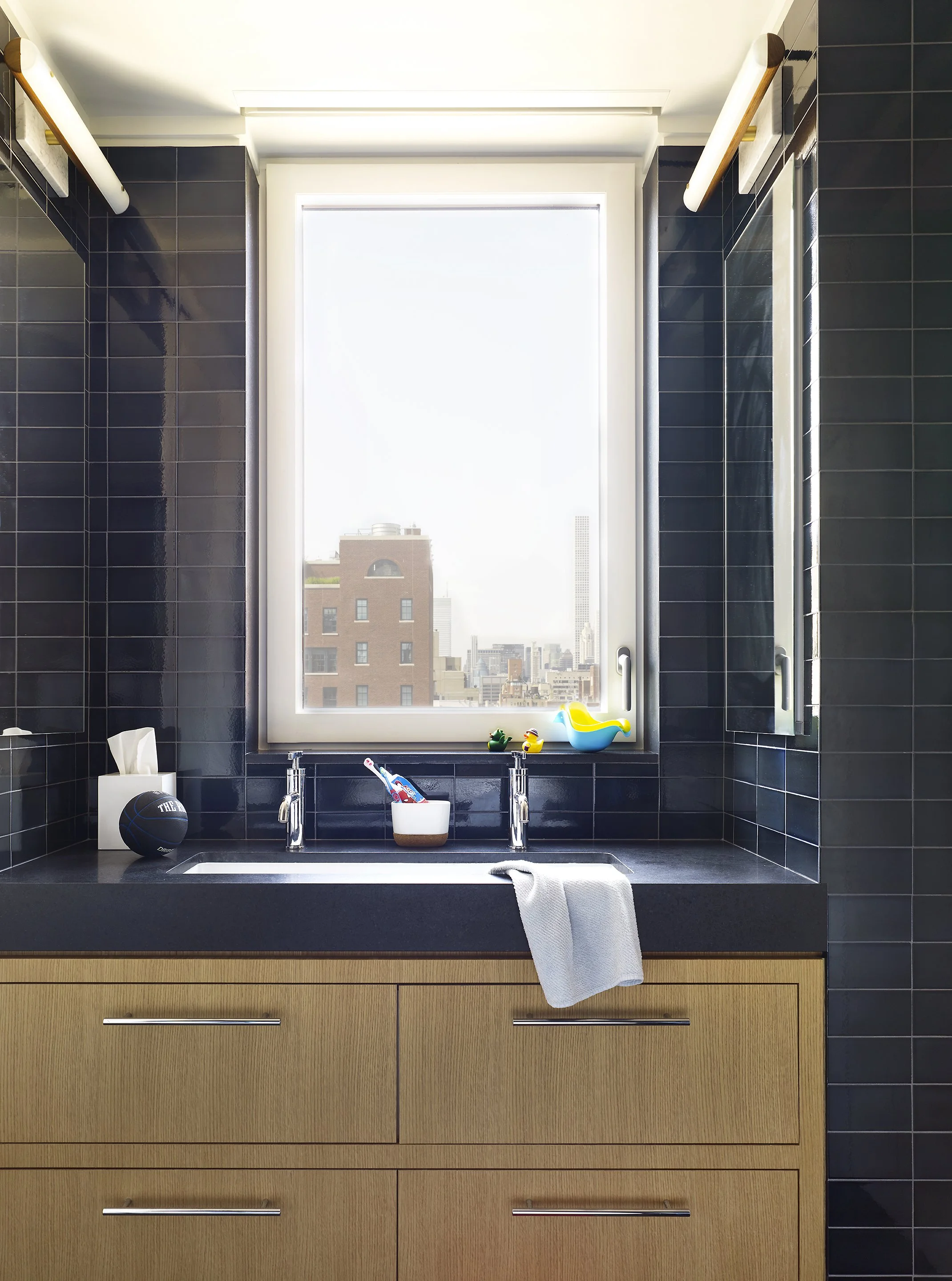 Modern bathroom with black tiles, a wooden vanity, a sink with a white towel, and a large window showing city buildings.